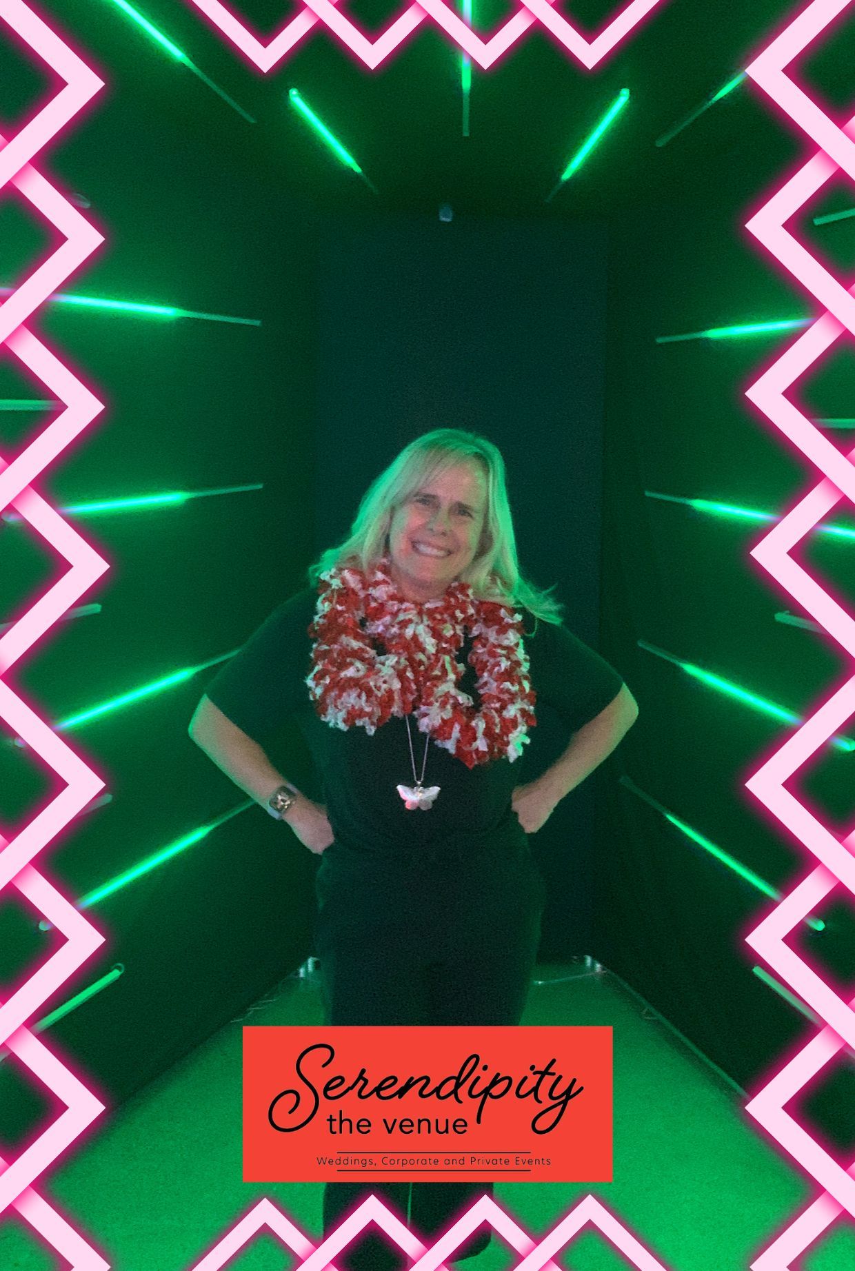 Woman standing in a neon-lit tunnel with pink zigzag borders, smiling and wearing a floral lei and black outfit
