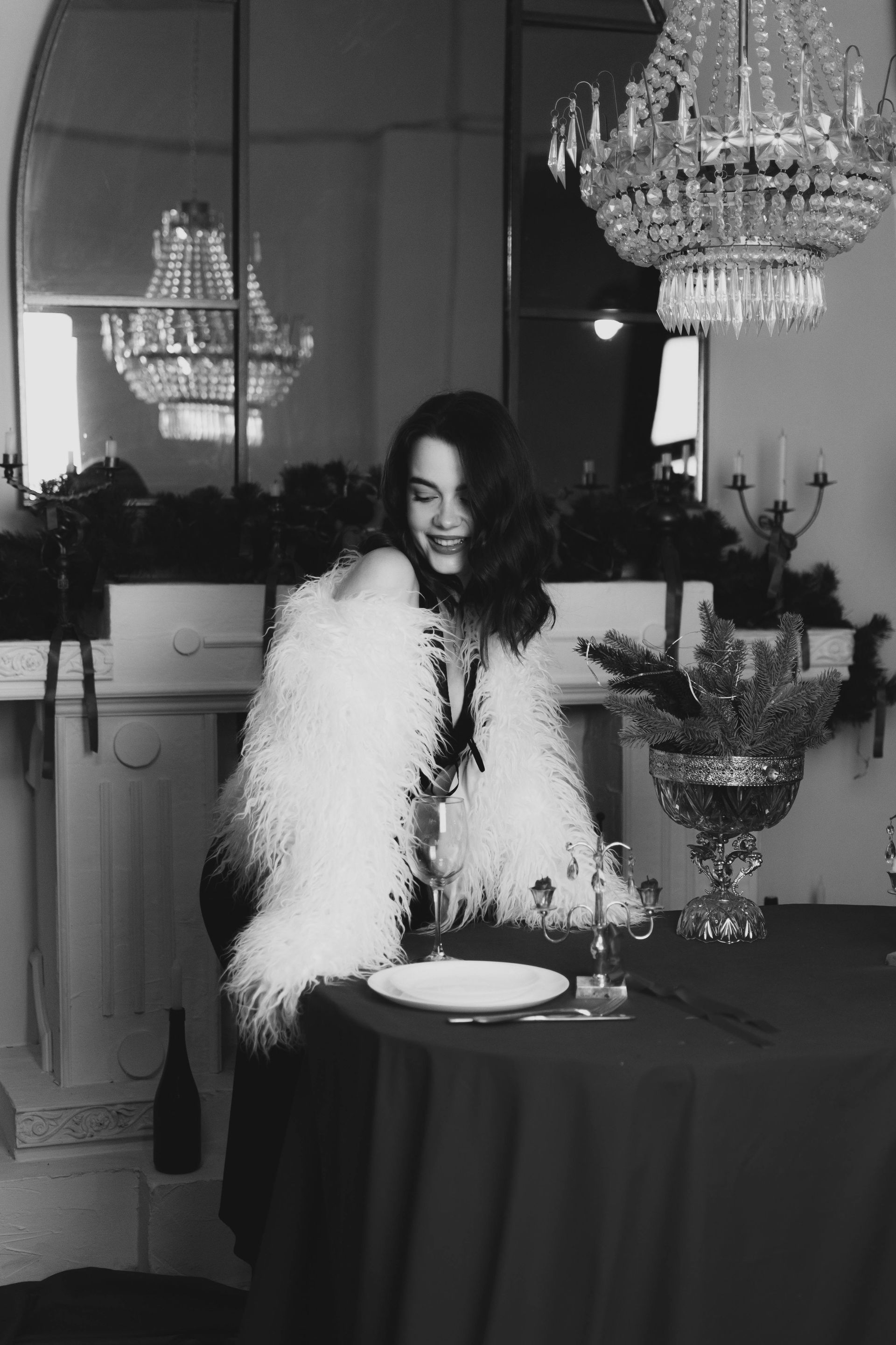 Woman in a feathered coat seated at a table in a chandelier-lit room, smiling with a rose arrangement nearby.