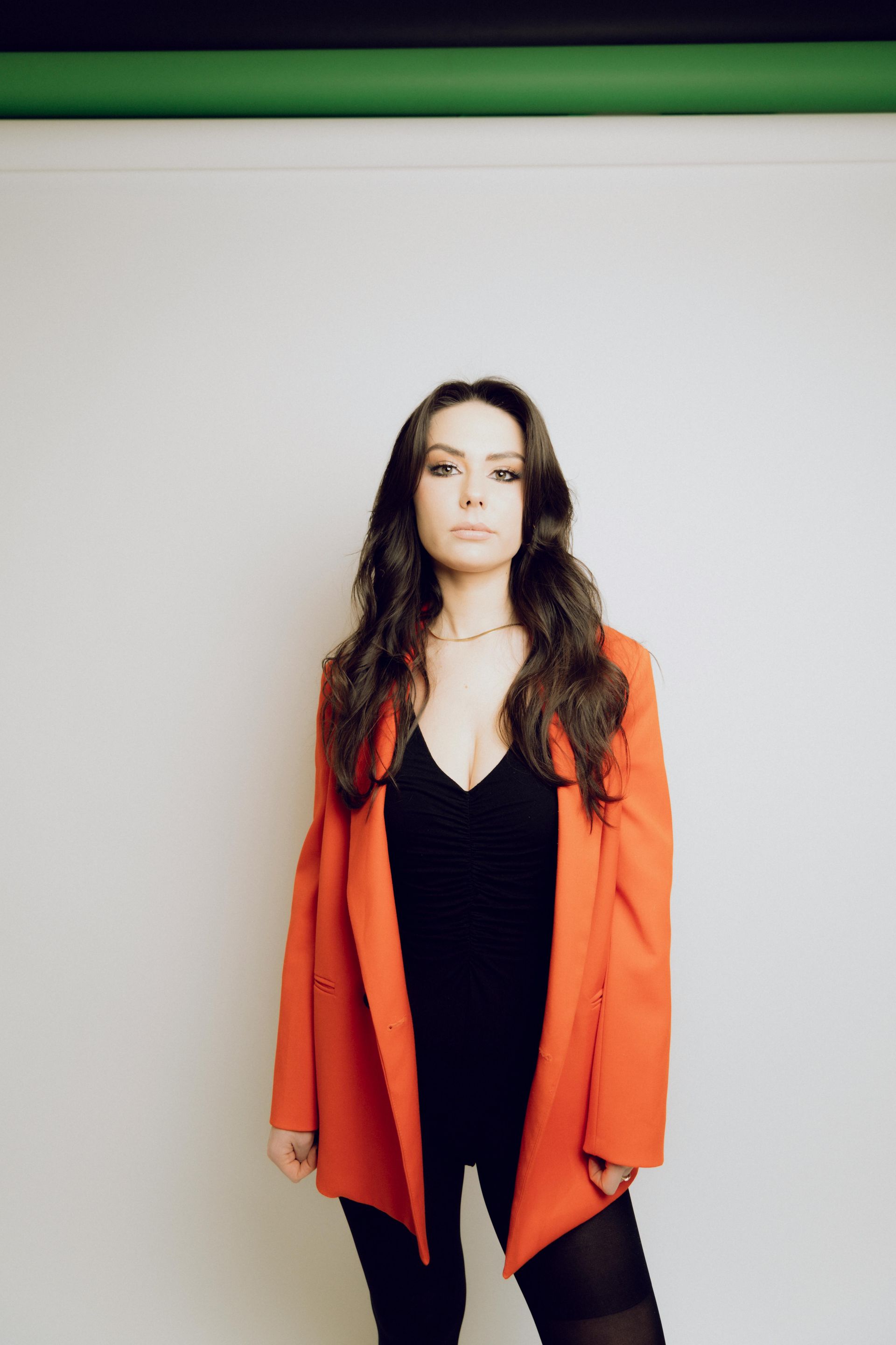 Woman standing against a white wall, wearing a bright orange blazer over a black outfit.