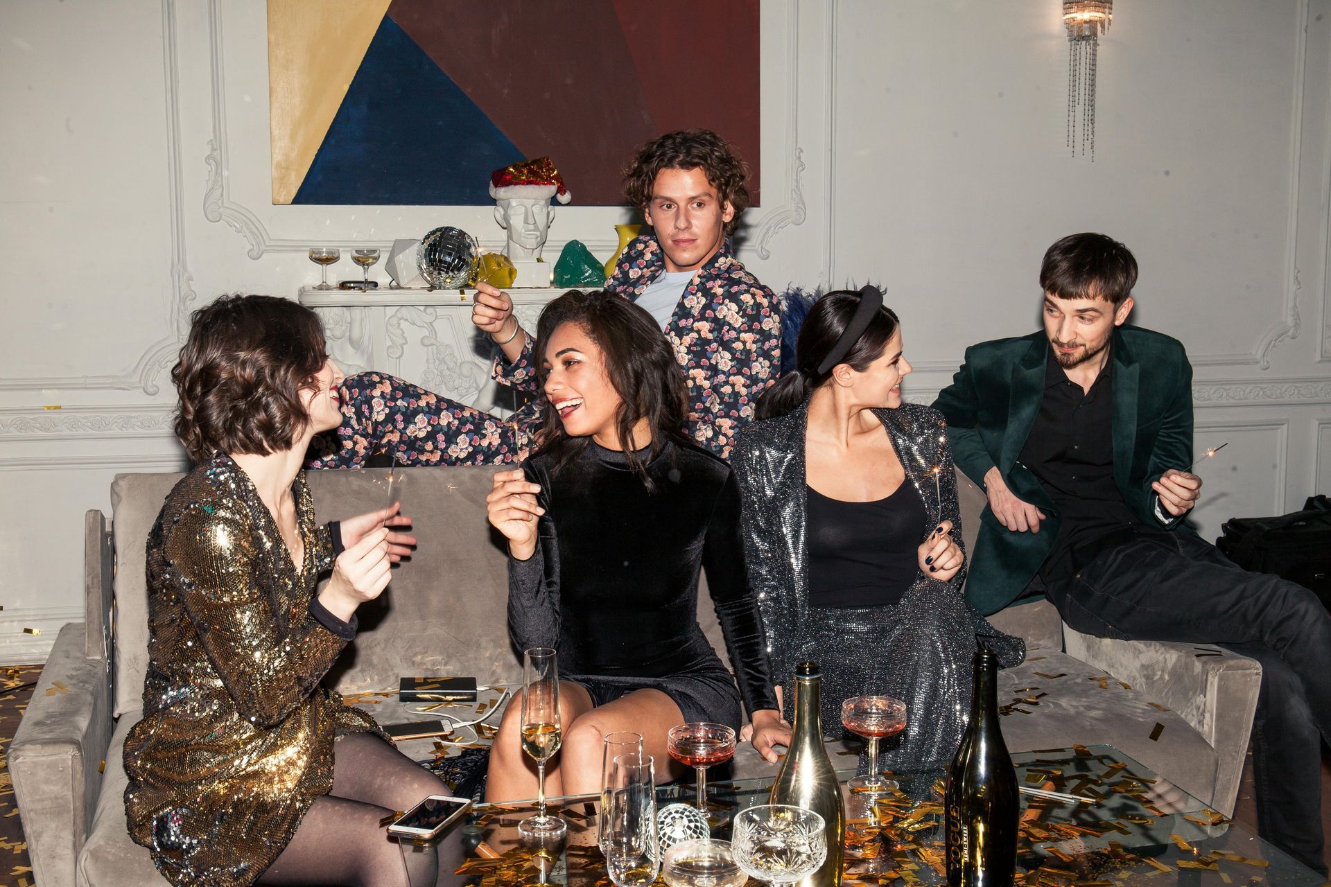 Five people chatting on a sofa at a stylish party, with drinks and a modern abstract painting behind them.