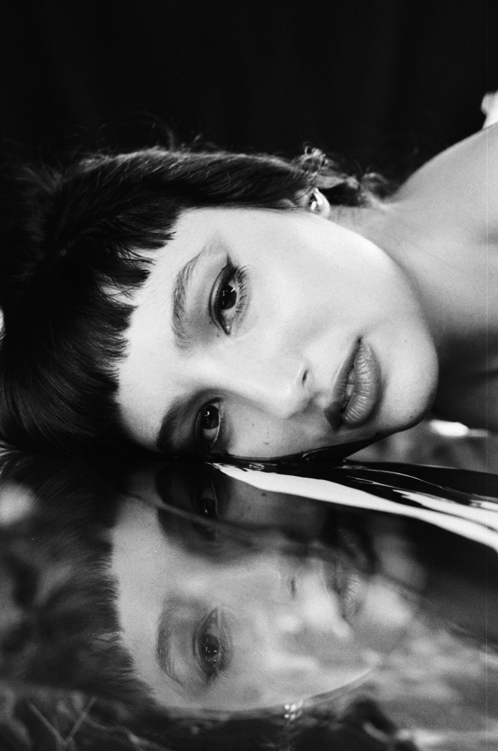Black-and-white close-up of a woman resting her head on a reflective surface, looking at the camera.