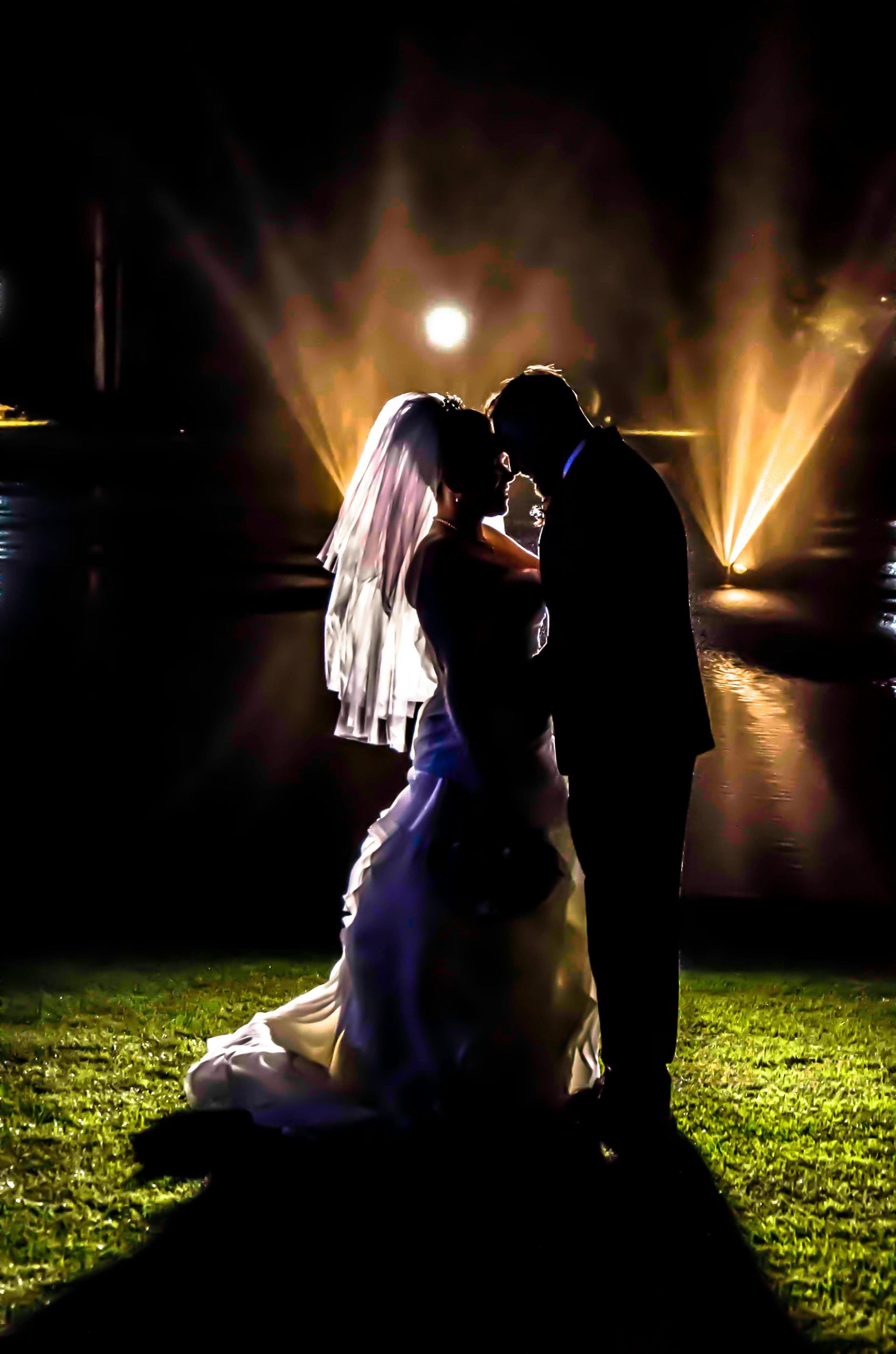Silhouetted couple embracing at night on a lit lawn, with glowing fountains behind them