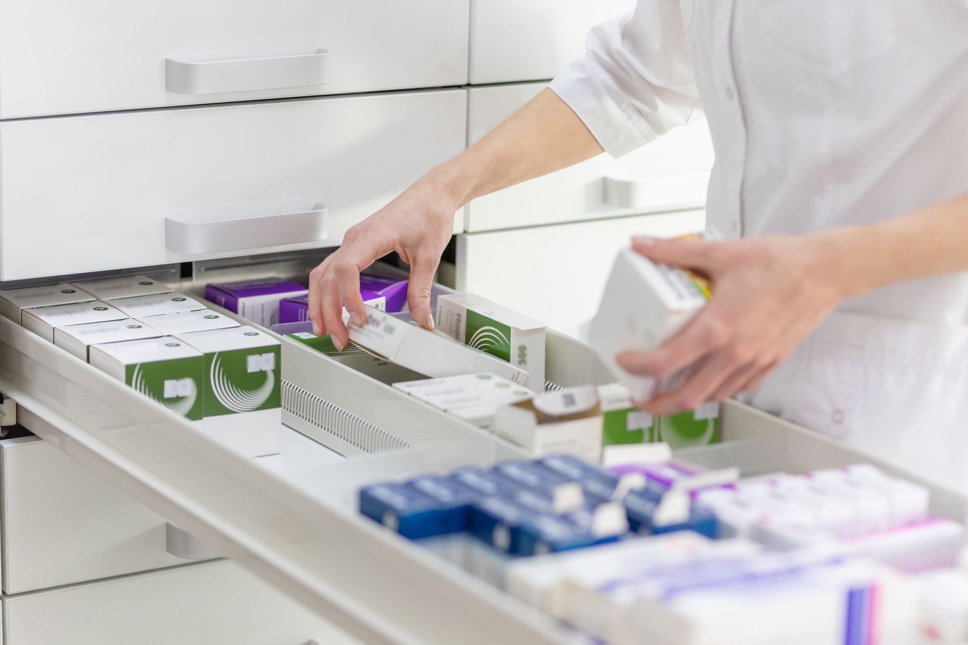 Eine Person legt Medikamentenpackungen in einen weißen Schubladenorganizer in einer hellen Apotheke oder Klinik.