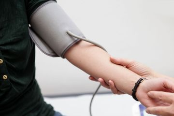 Eine Blutdruckmanschette ist am Arm einer Person angelegt, während eine andere Person den Puls prüft.