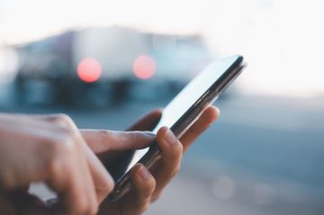 Hände, die ein Smartphone benutzen; im Hintergrund sind ein verschwommenes Auto und Straßenlaternen zu sehen.