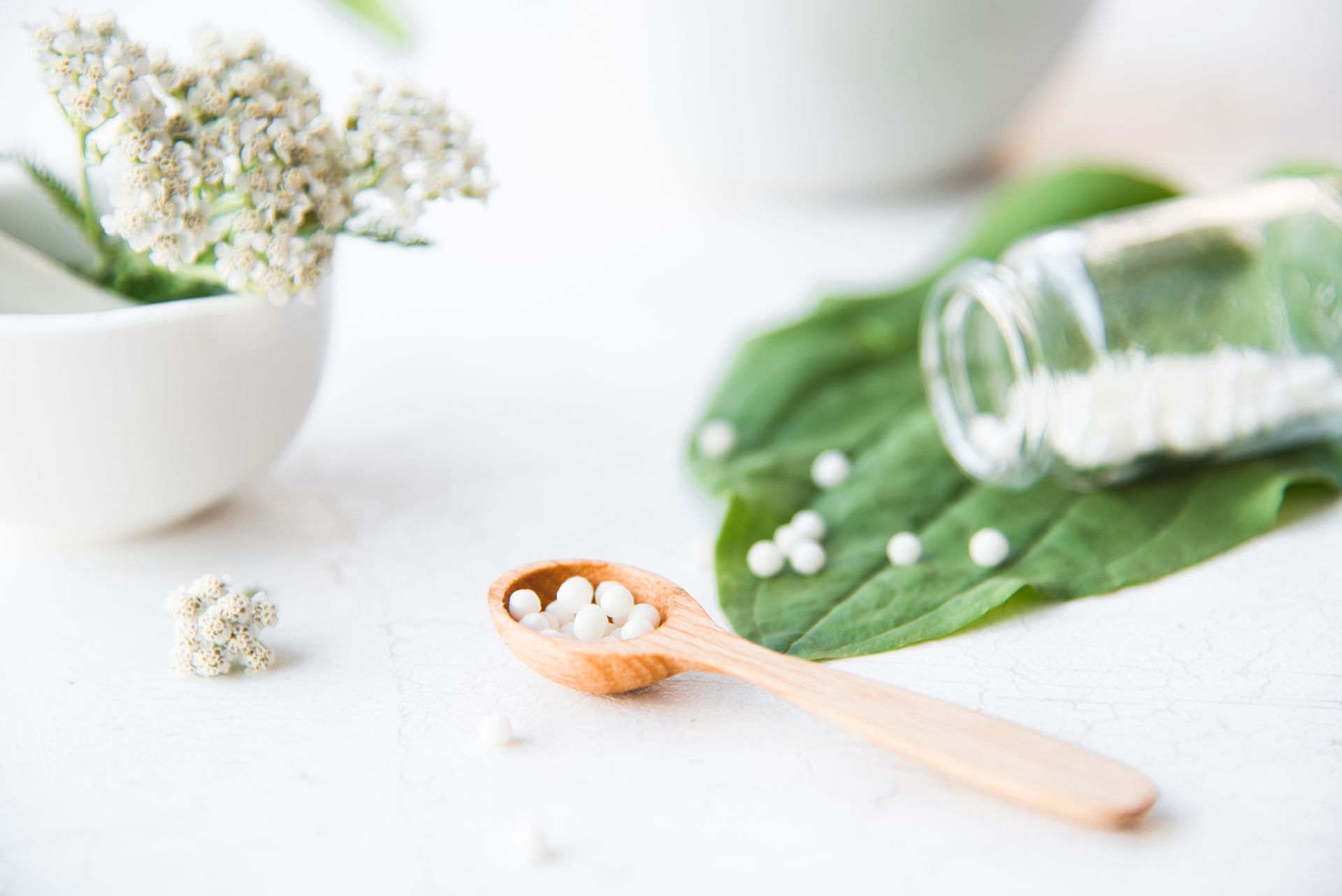 Weißer Löffel mit Tabletten neben einem umgekippten Glasgefäß und einem grünen Blatt auf weißem Grund