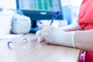 Behandschuhte Hände entnehmen Blut in ein Reagenzglas an einem Klinikschreibtisch