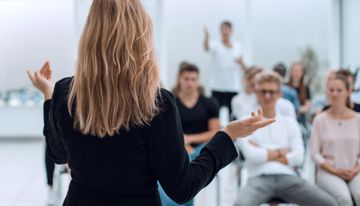 Ein/e Moderator/in spricht zu einem Publikum in einem hellen Klassenzimmer oder einer Werkstatt.