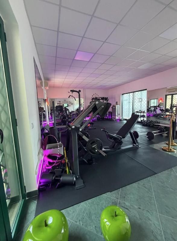 Interno di una palestra con macchine per esercizi, specchi, illuminazione viola e kettlebell verdi in primo piano.