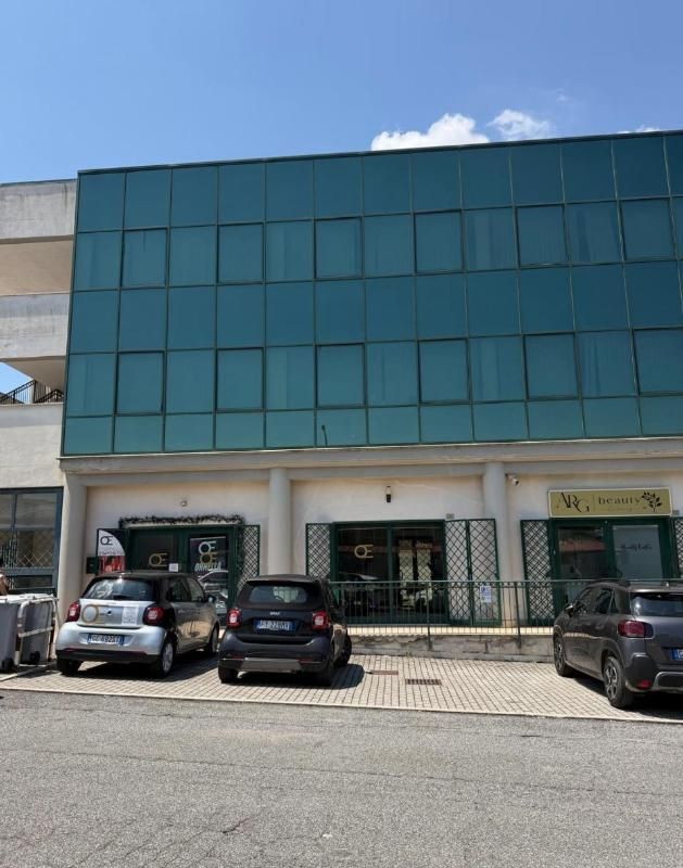 Vetrina moderna con facciata in vetro color verde acqua, auto parcheggiate e un ingresso con cancello sotto un cielo azzurro limpido.