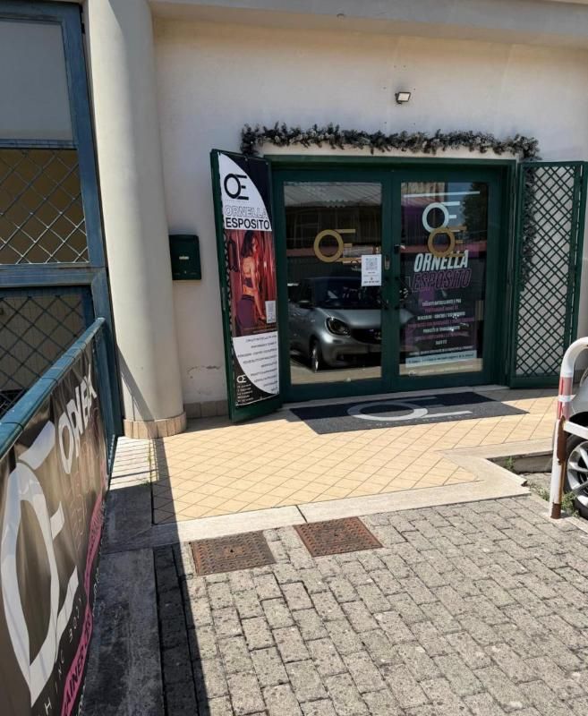 Ingresso del negozio con porte a vetri, passaggio piastrellato e cancello laterale in rete verde in una giornata di sole
