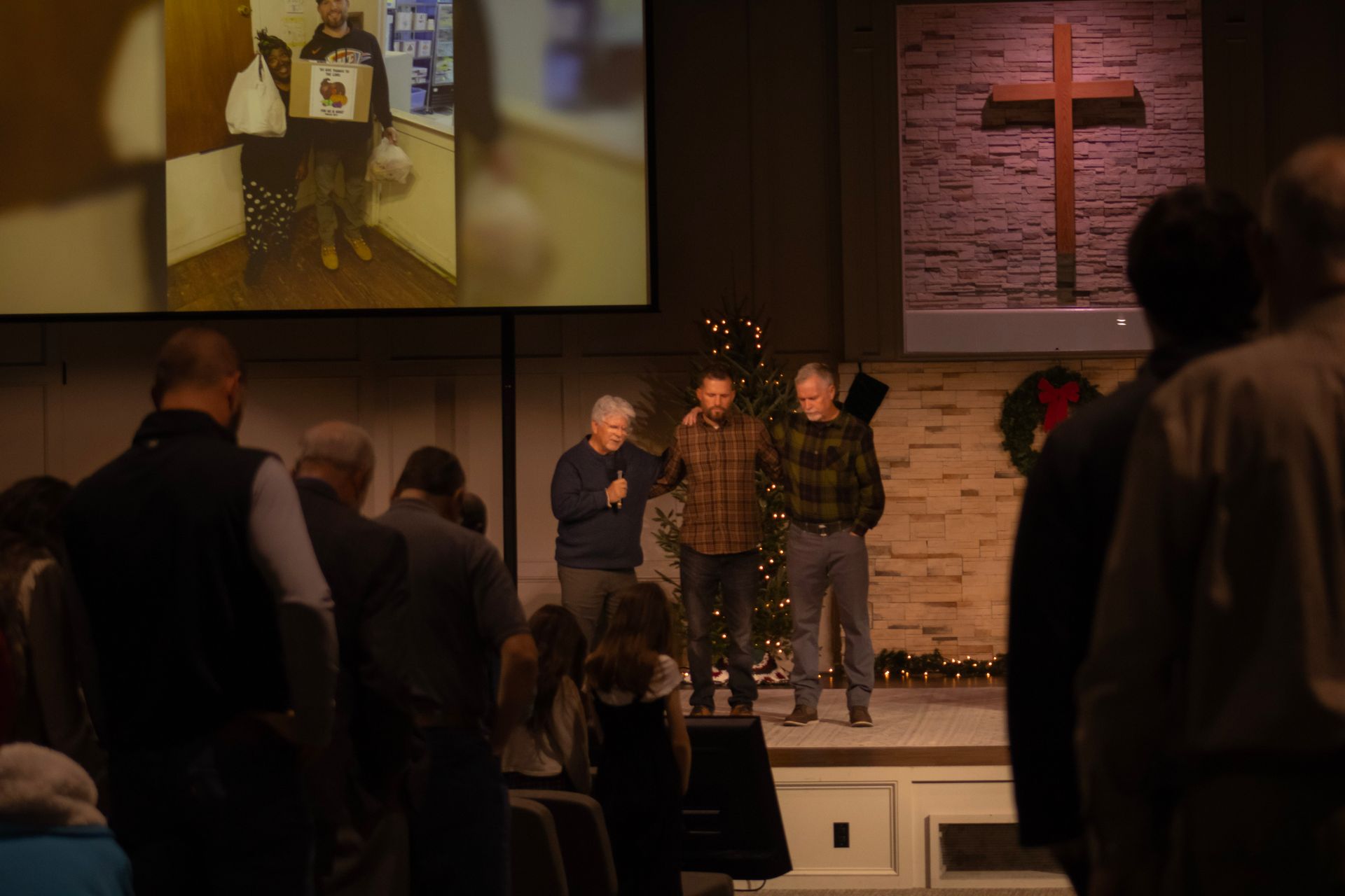 Three individuals stand on a church stage during a service, with a large screen displaying a photo above them.