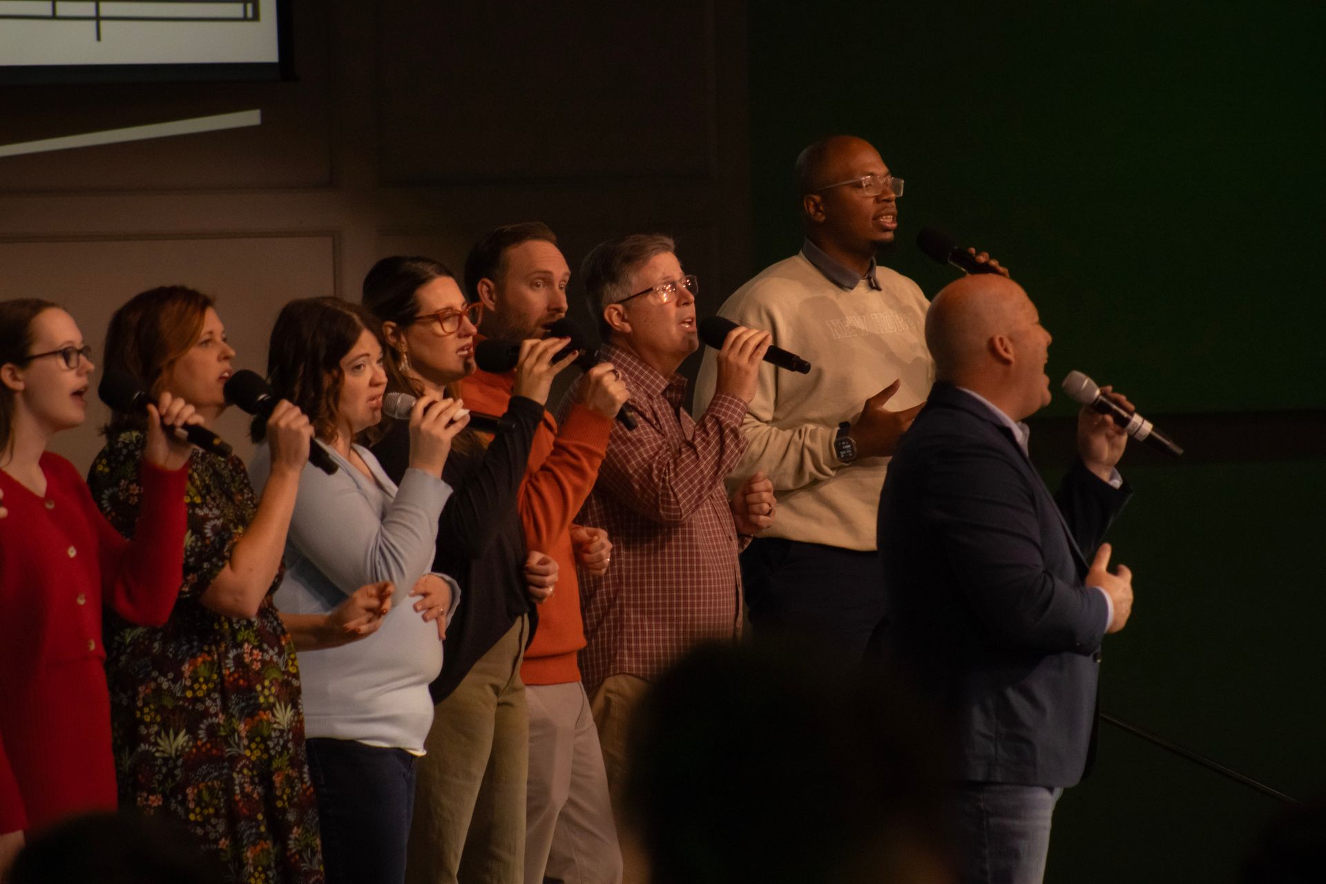 A group of people standing on a stage, singing into microphones in a dimly lit indoor setting.