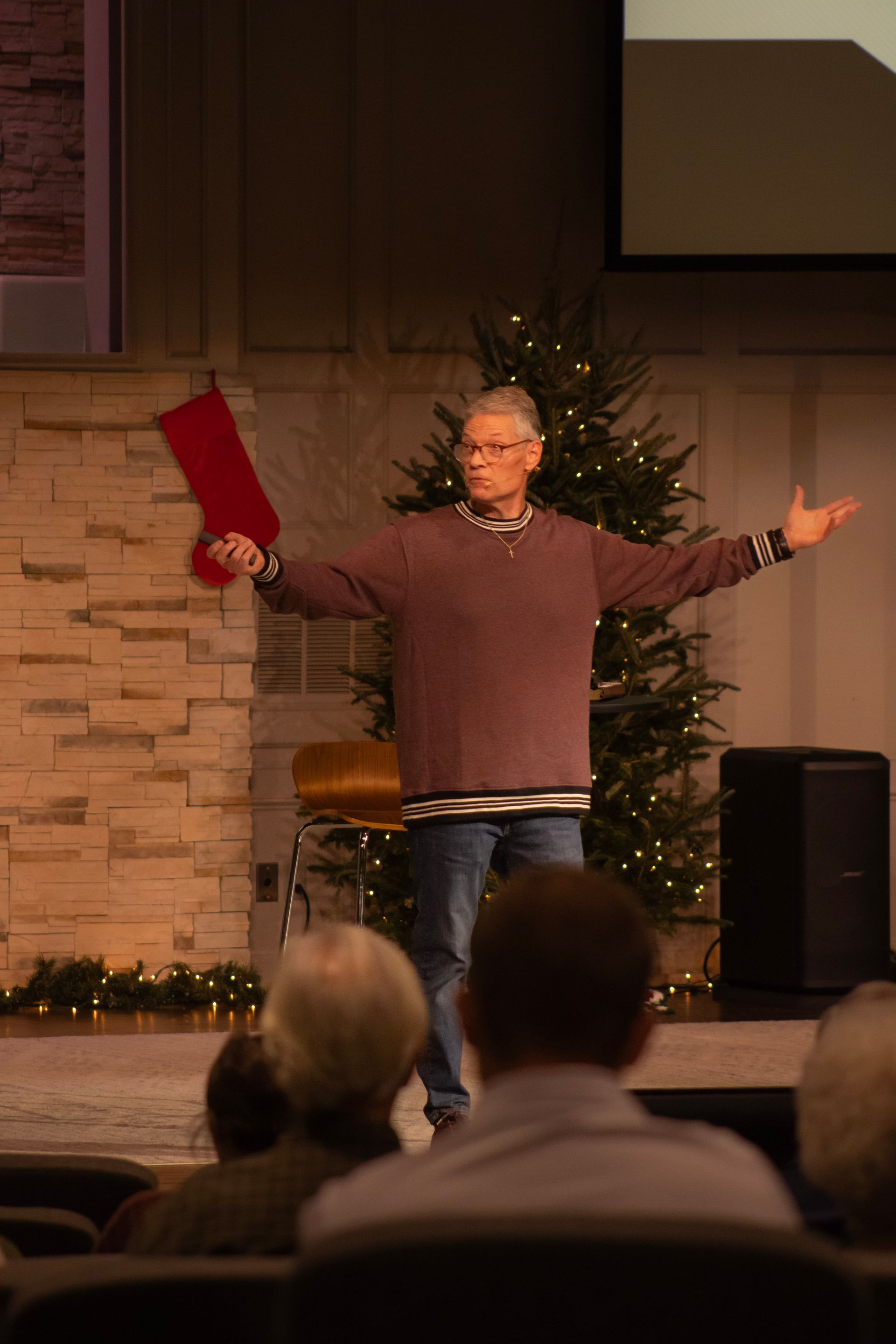 A speaker stands on a stage with arms outstretched, in front of a Christmas tree and a stone-textured wall.