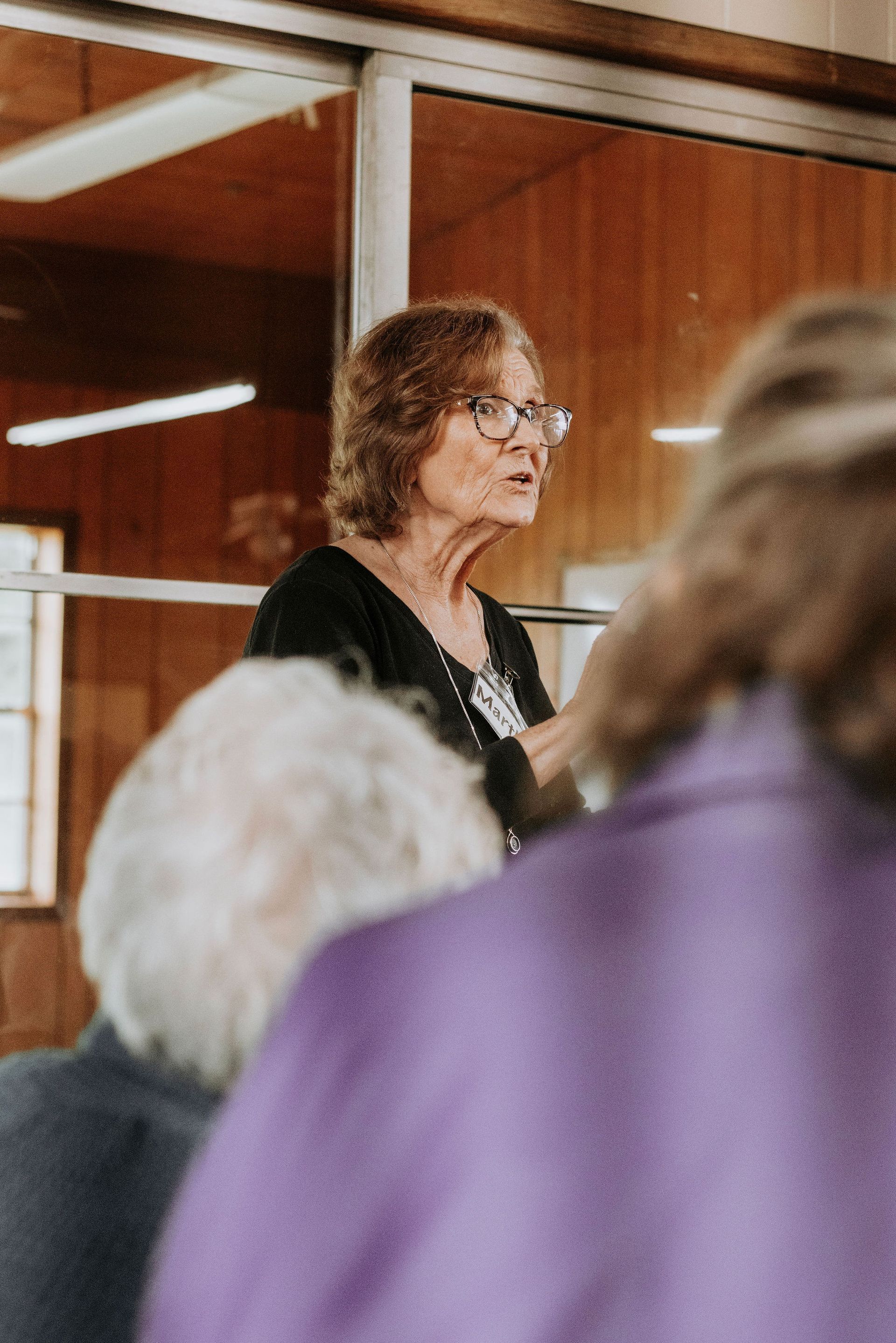 Woman with glasses speaking at an event, wooden panel background.