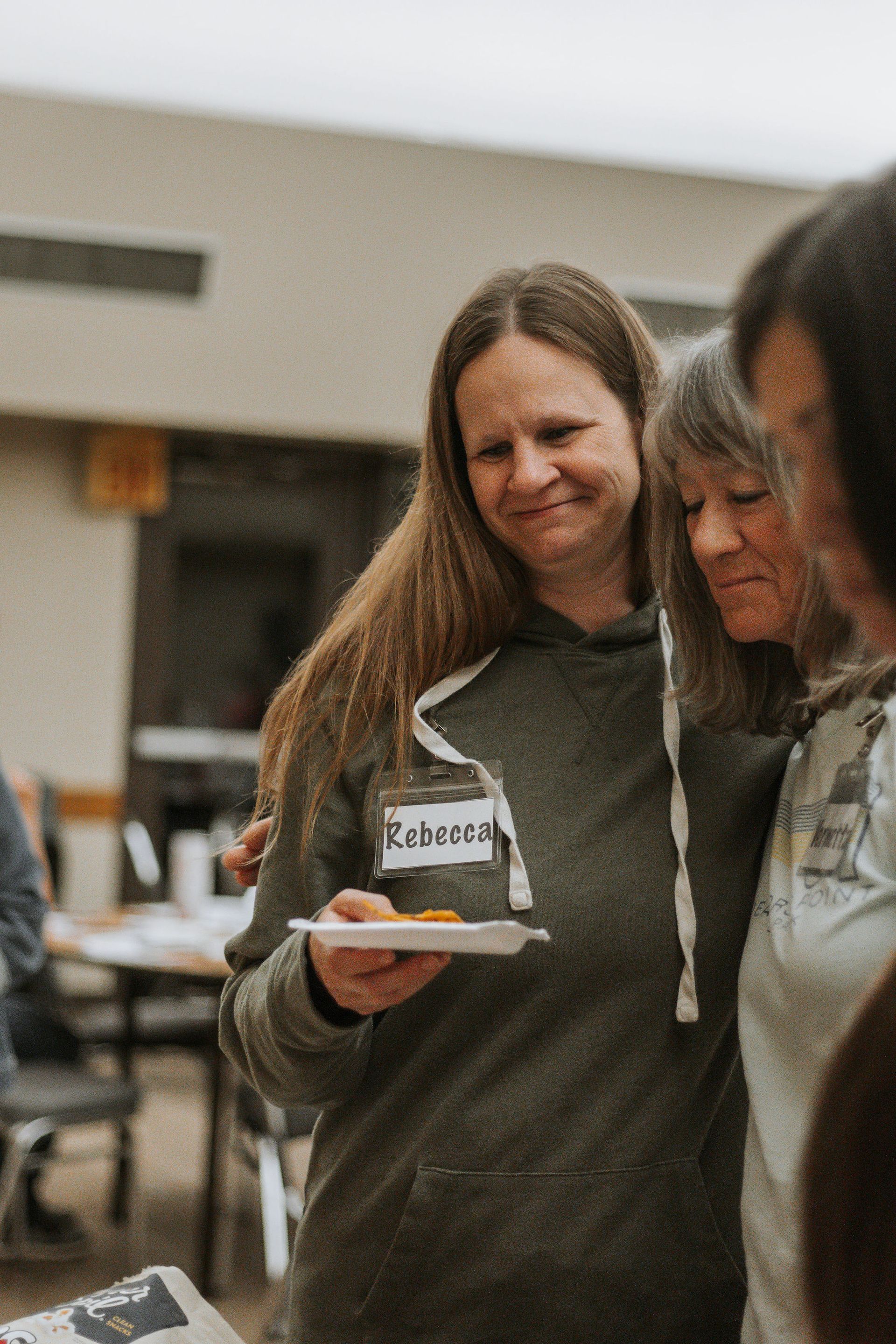 Woman with long hair holds plate, smiles near older woman with gray hair; indoors.