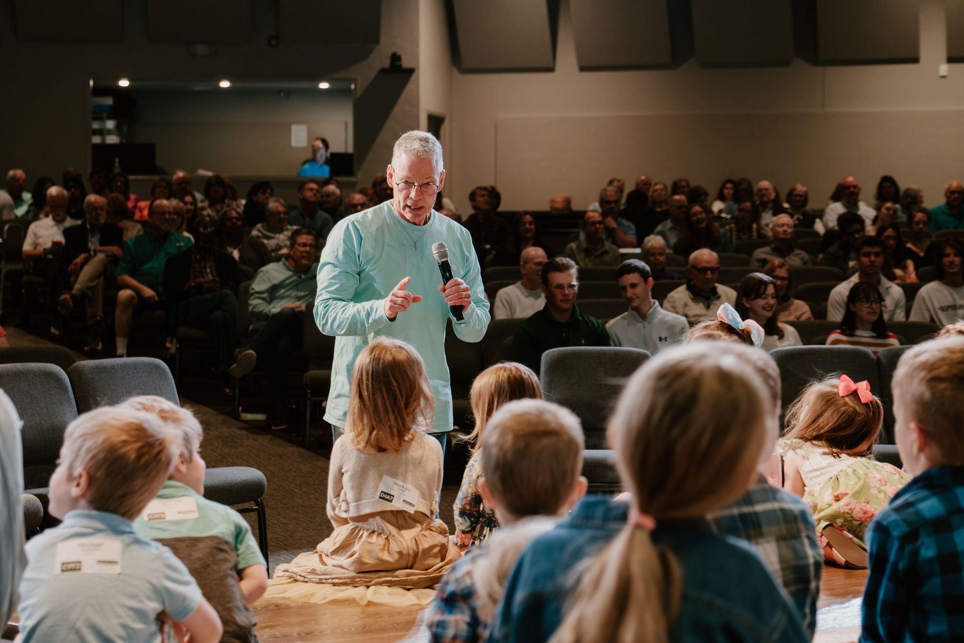 Speaker in a teal shirt addressing a seated audience in a large hall