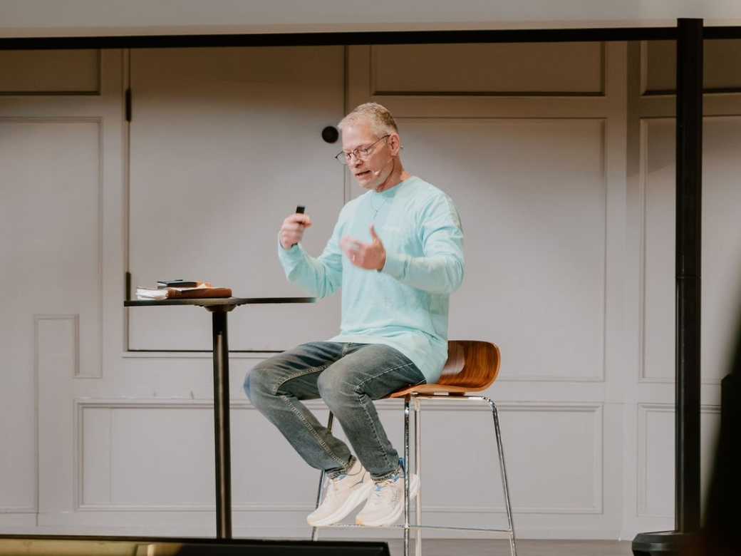 Man speaking into a headset microphone while seated on a stool beside a small table in a simple studio set