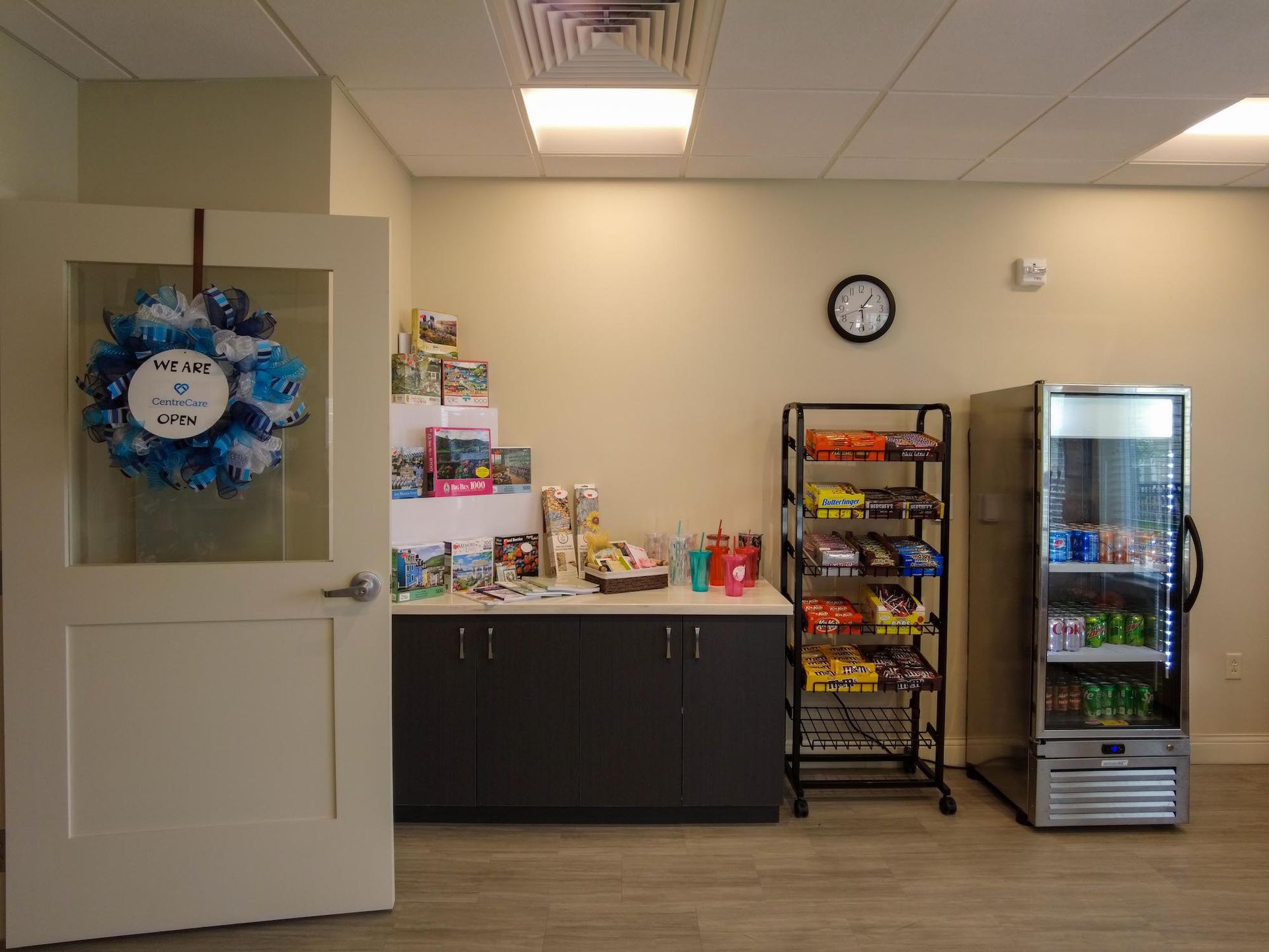 Room with a door, snacks, and a vending machine. A clock is on the wall. A blue wreath hangs on the door.