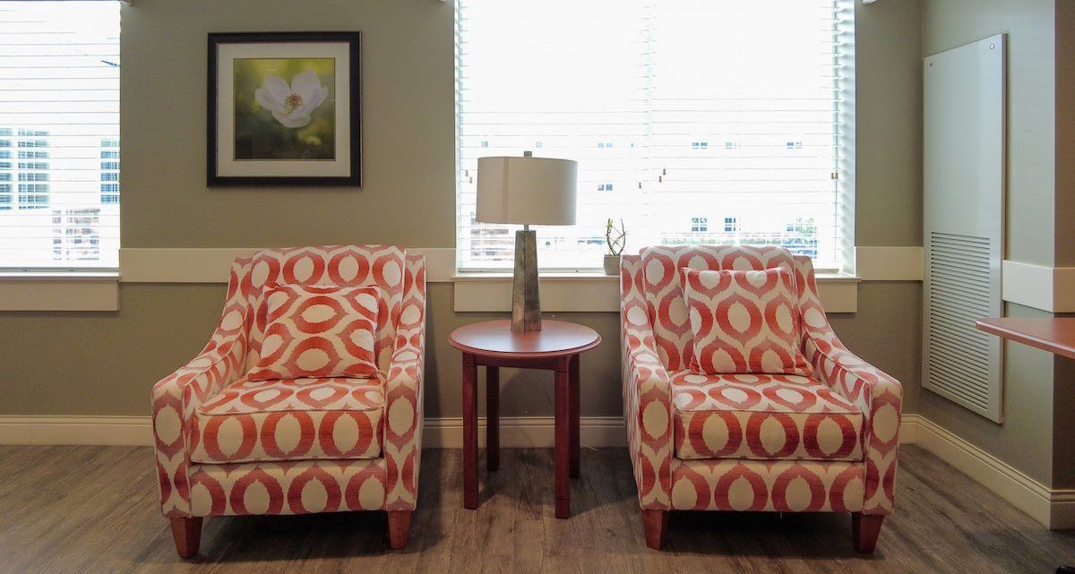 Two patterned armchairs and a side table with a lamp sit between two windows. A framed picture hangs above the chairs.