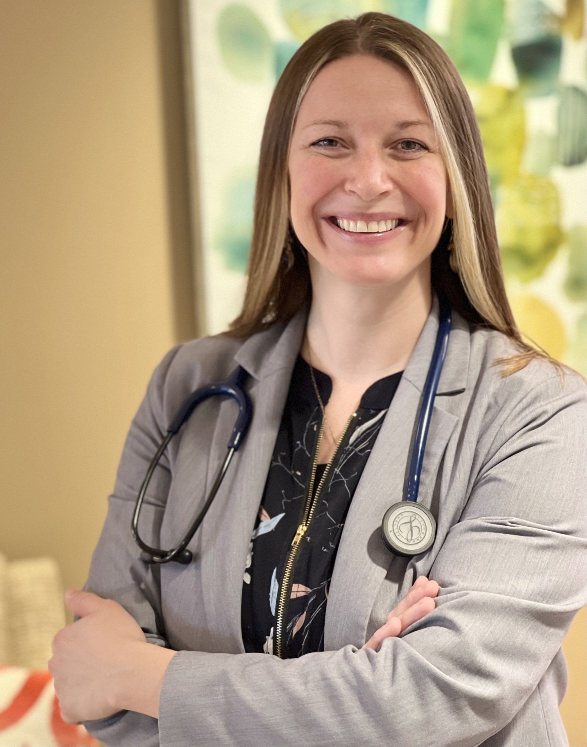 Xenia, CRNP wearing a gray blazer with stethoscope smiles, arms crossed; indoor setting.