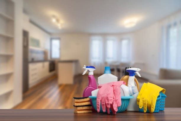 Cleaning supplies, pink and yellow gloves, and sponges on a table in front of a blurred living room.