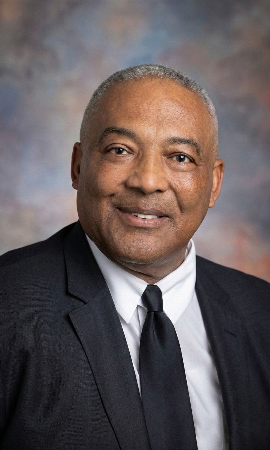 Portrait of a smiling man in a dark suit and tie against a gray-blue studio background