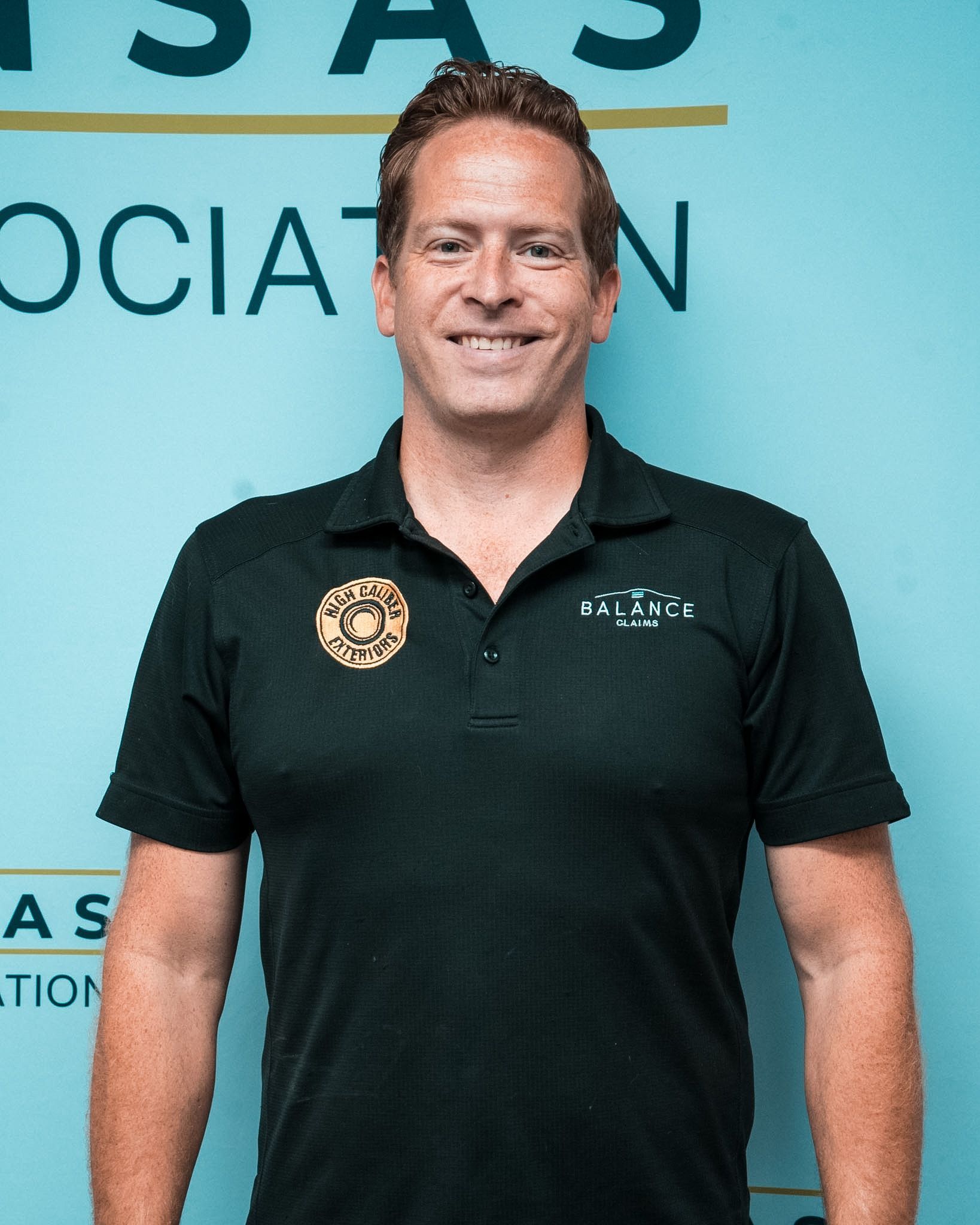 Man in black polo shirt standing at an event backdrop with teal signage