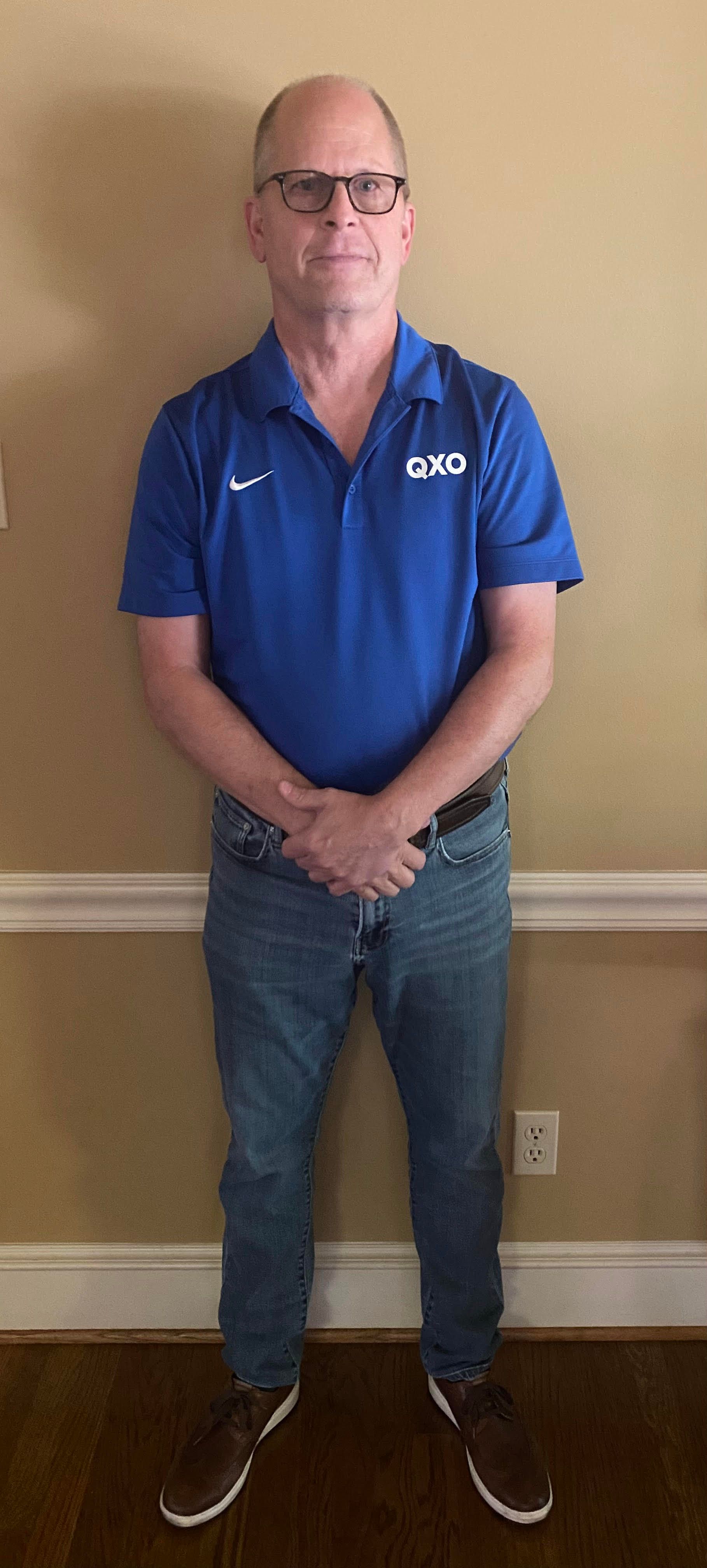 Man standing indoors against a plain wall, wearing a blue polo, jeans, and glasses.