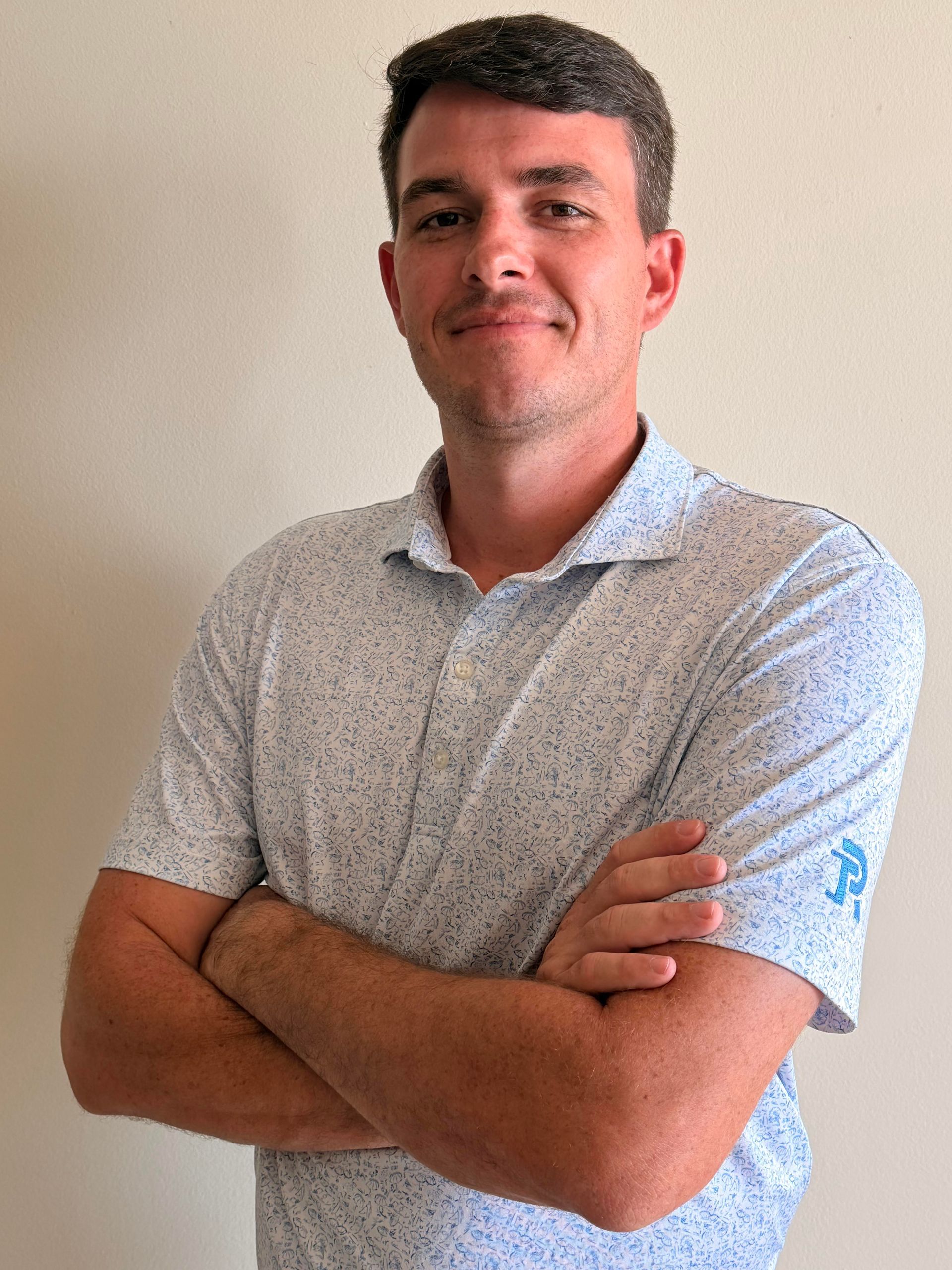 Man standing with arms crossed, wearing a light patterned shirt against a plain wall.
