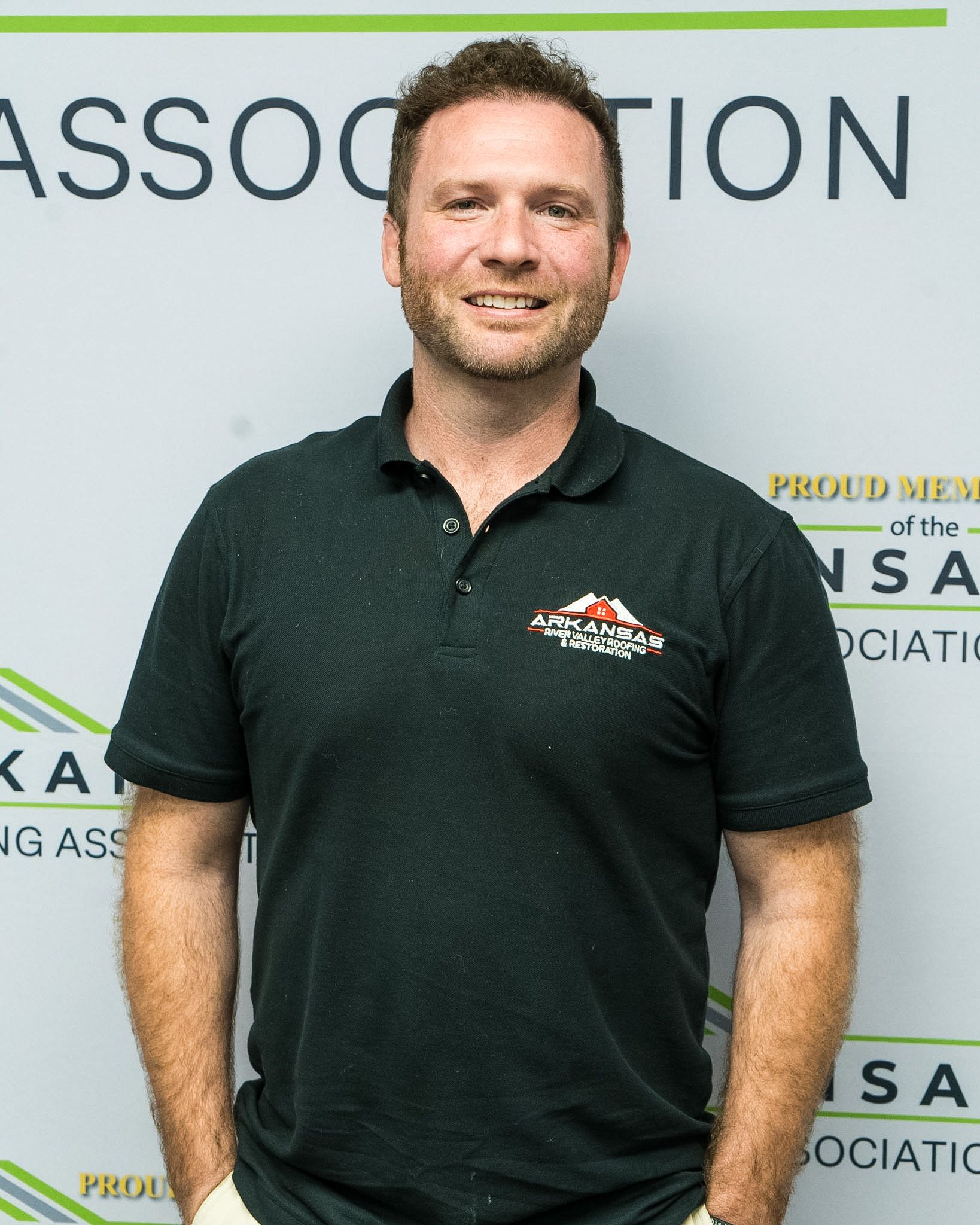 Man standing in front of an association backdrop, wearing a dark green polo shirt and smiling.