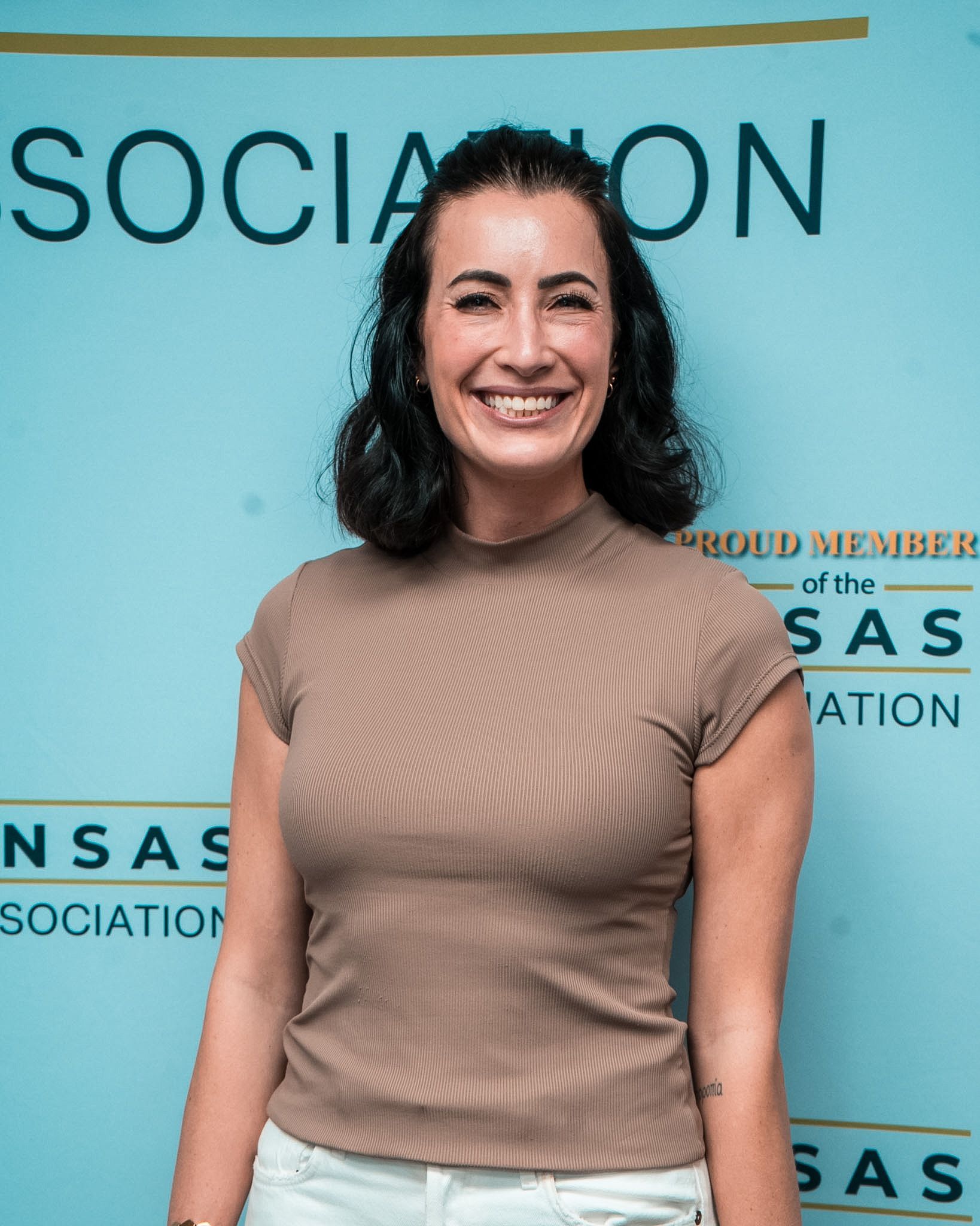 Smiling woman in a tan top standing before a turquoise event backdrop with text
