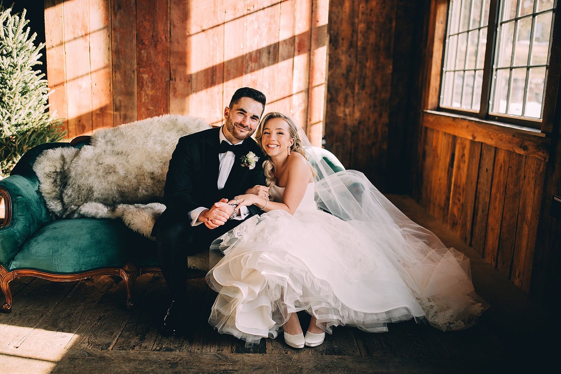 Newlyweds seated on a teal couch, bride in a wedding dress, groom in a tuxedo, smiling, rustic setting.