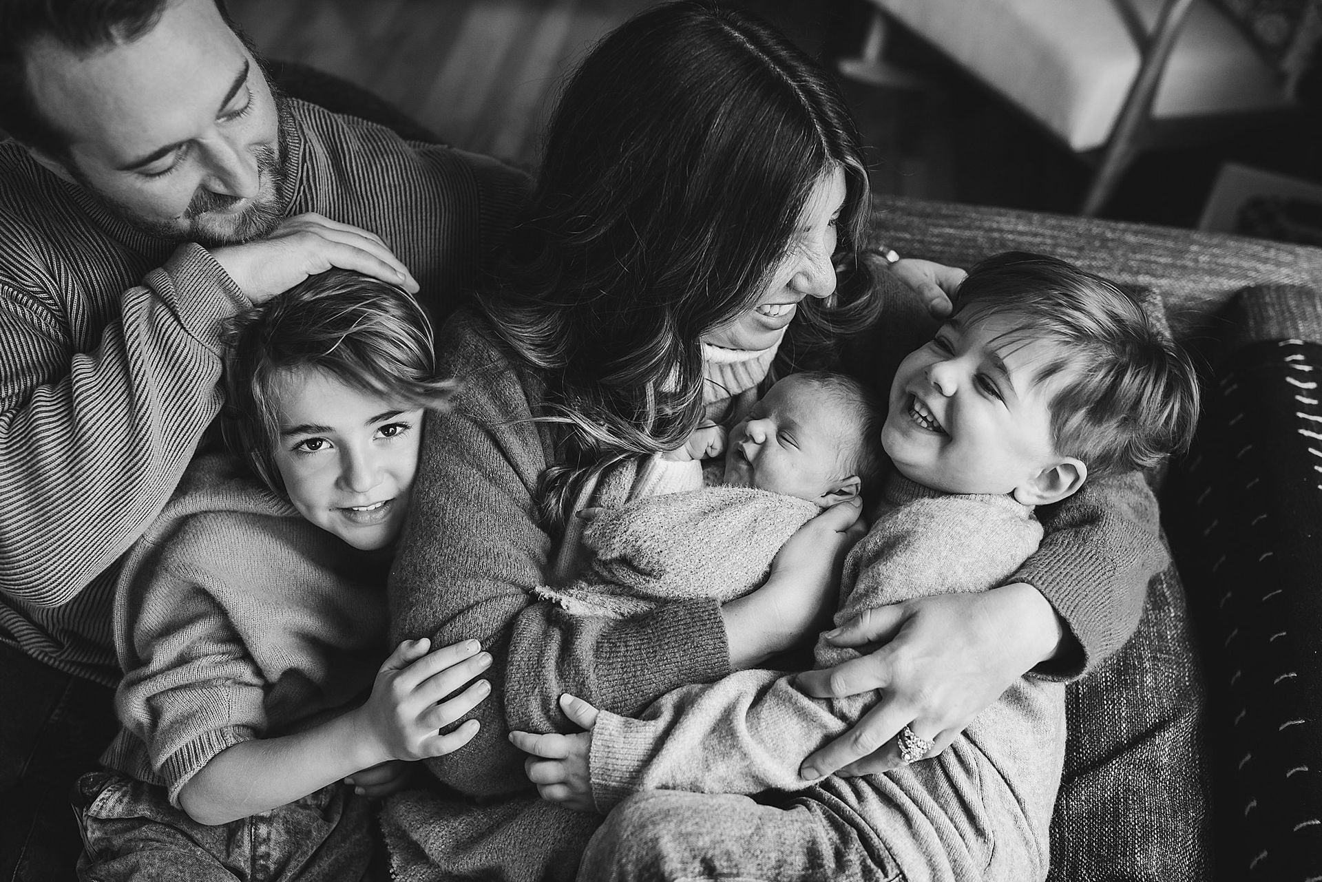 Family portrait: mother holding newborn and toddler, older child looks at camera, father smiles.