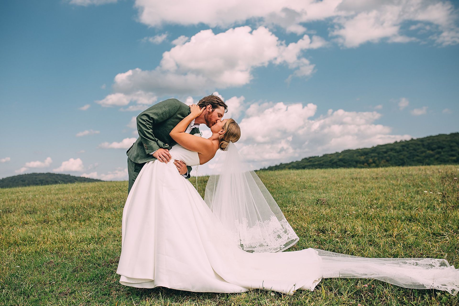 Couple kissing in a field on their wedding day; bride in white dress, groom in suit; sunny sky, mountains in background.