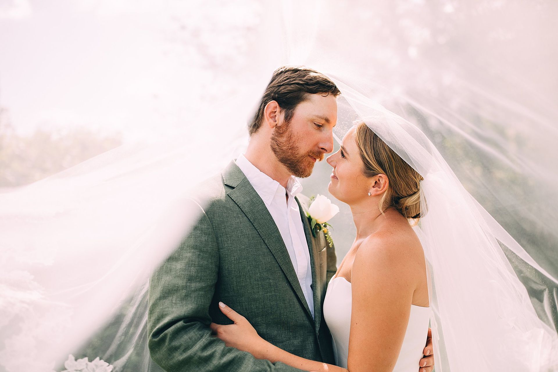 Bride and groom embrace under a veil outdoors, faces close, soft lighting.