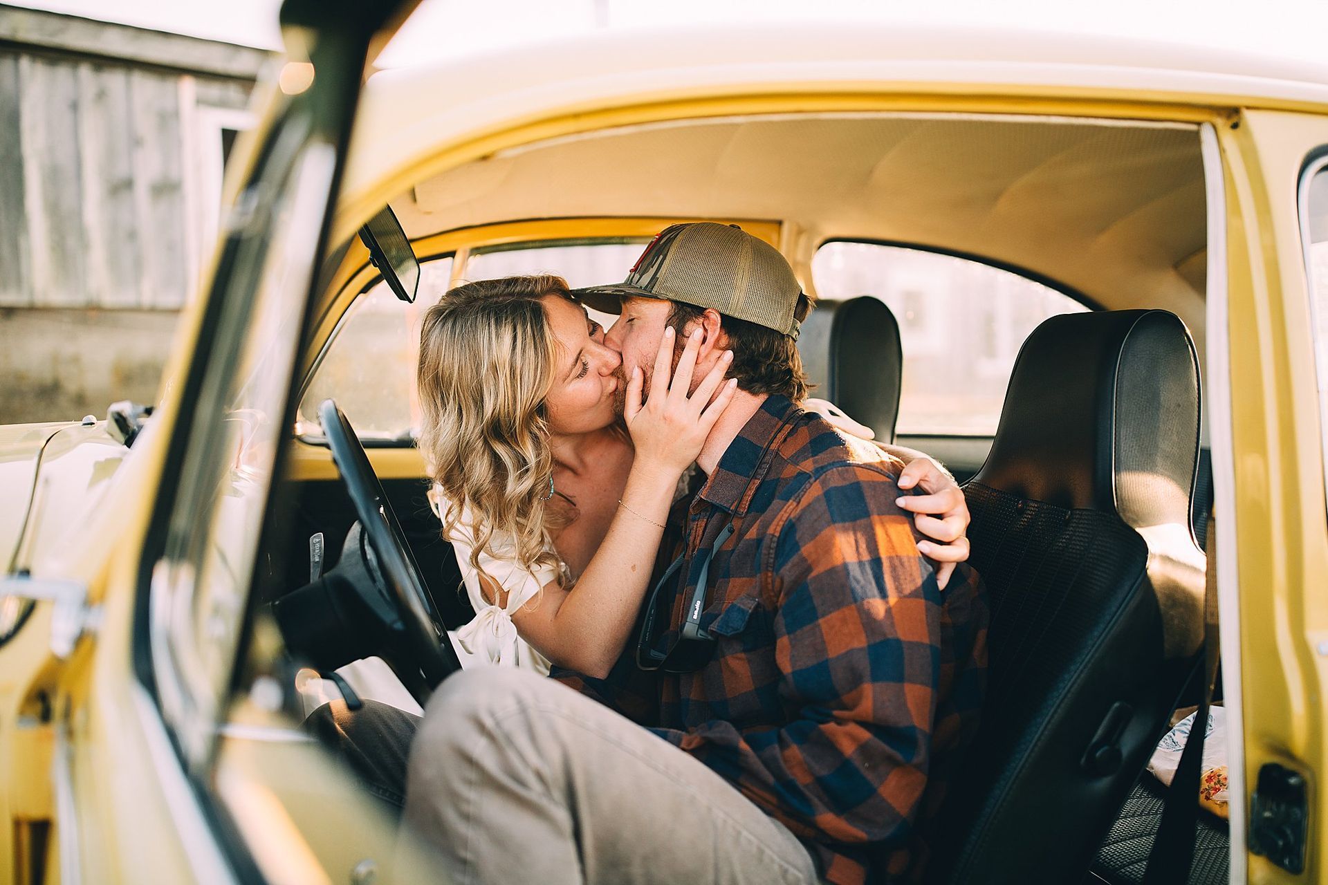 Couple kissing inside a vintage yellow car; he wears a plaid shirt and hat, she has curly hair.