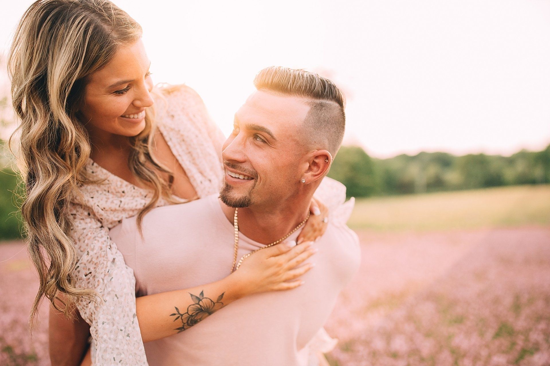 Woman smiles, sits on man's back, in a field of lavender. Man grins, looking at woman.