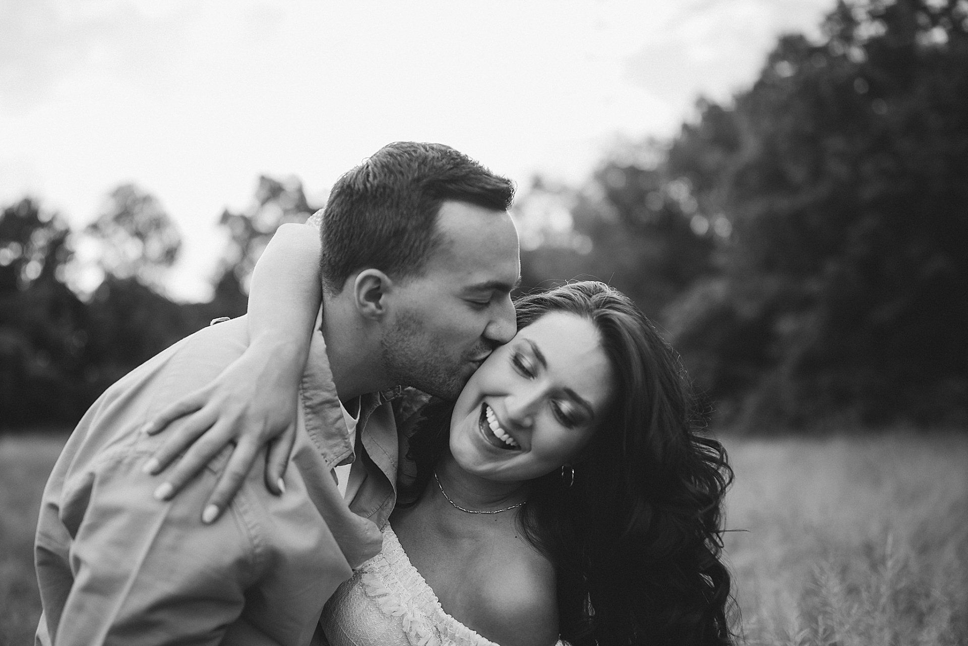 Man kisses woman's cheek in a field, both smiling. Black and white photo.