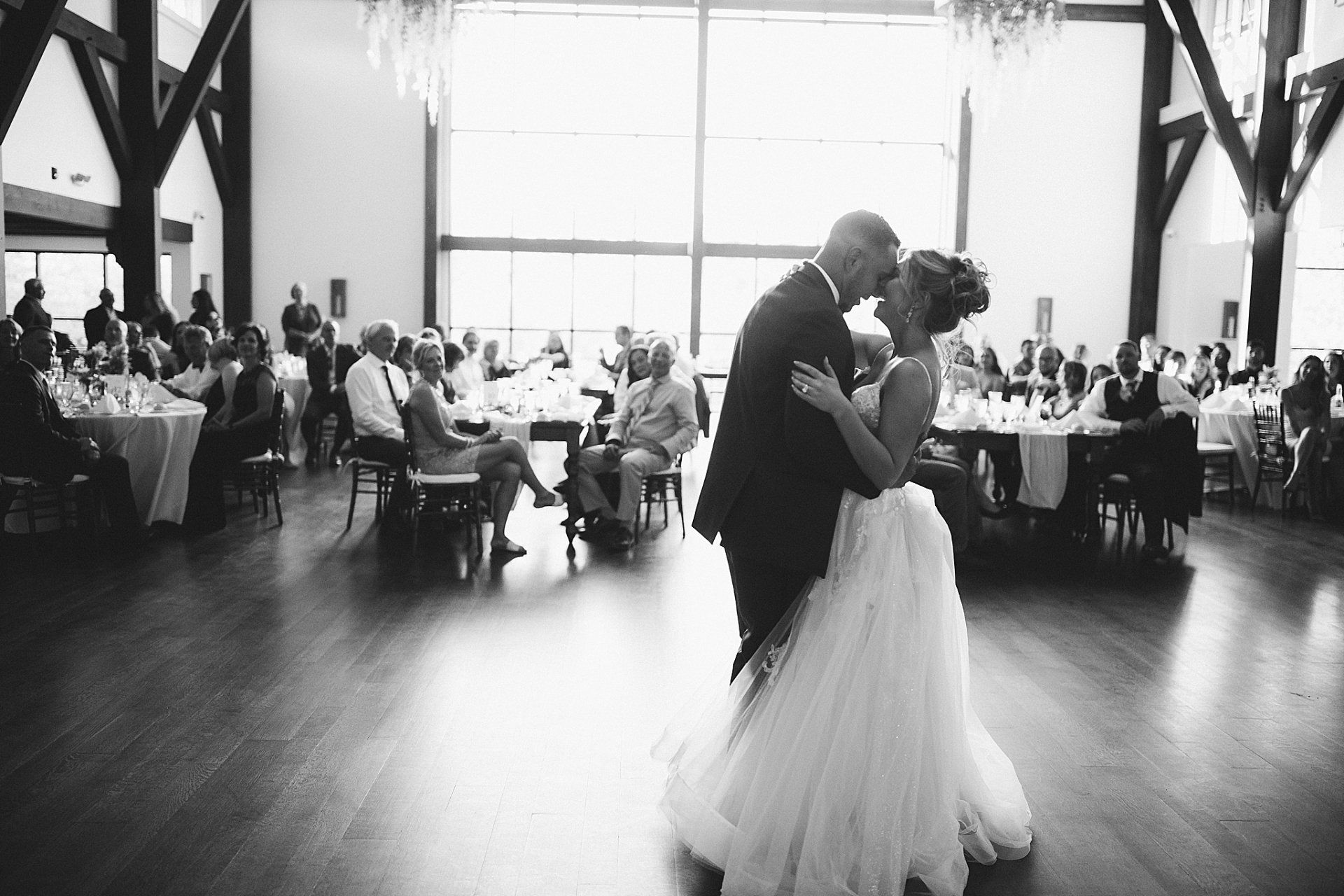 Bride and groom dance at reception; guests seated at tables in a large hall with windows. Black and white.