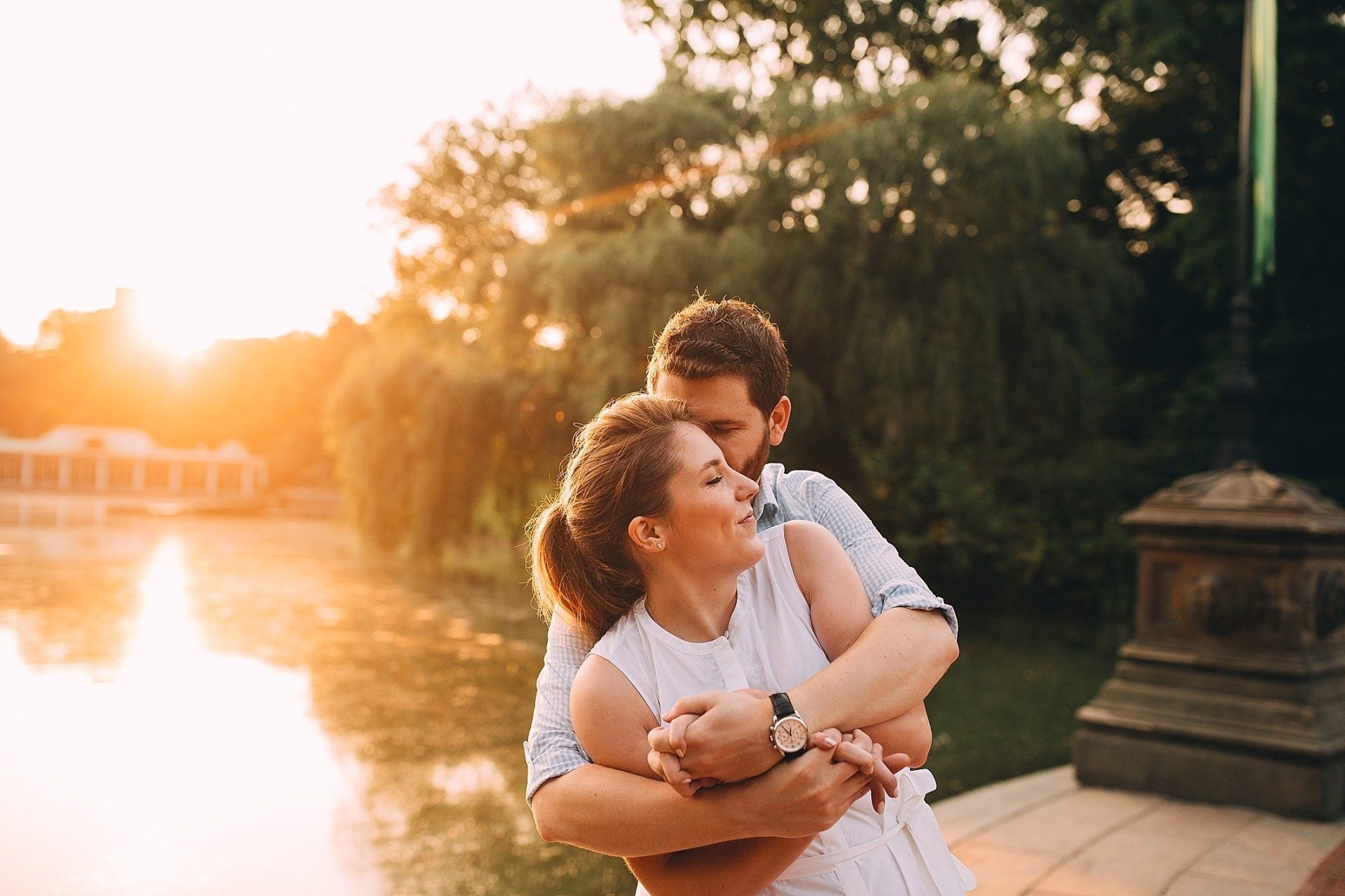 Couple embracing by a lake at sunset. Man's arms around woman, both smiling, bright golden light.
