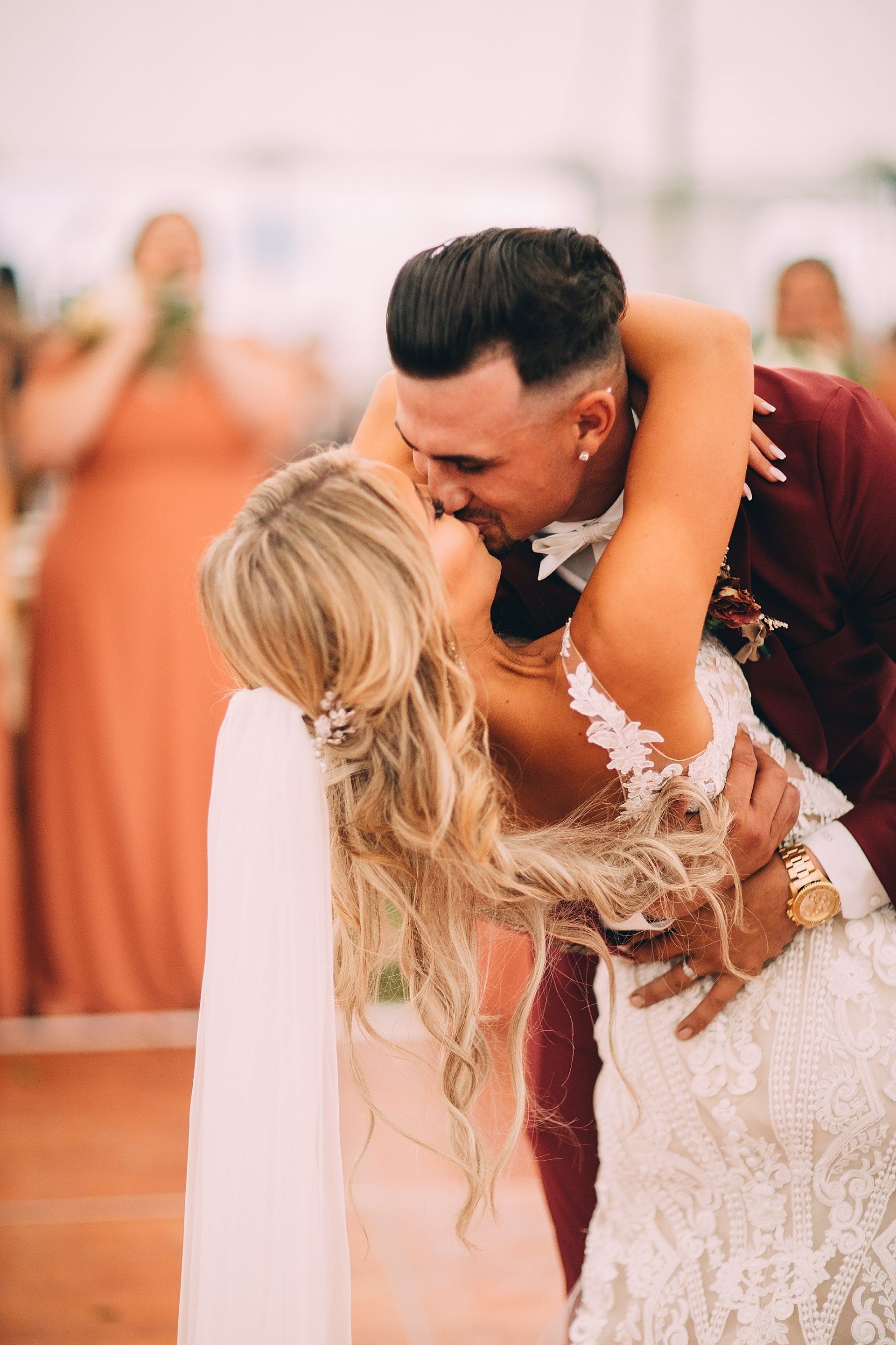 Bride and groom embrace, kissing during wedding reception; she in white lace dress, he in burgundy suit.