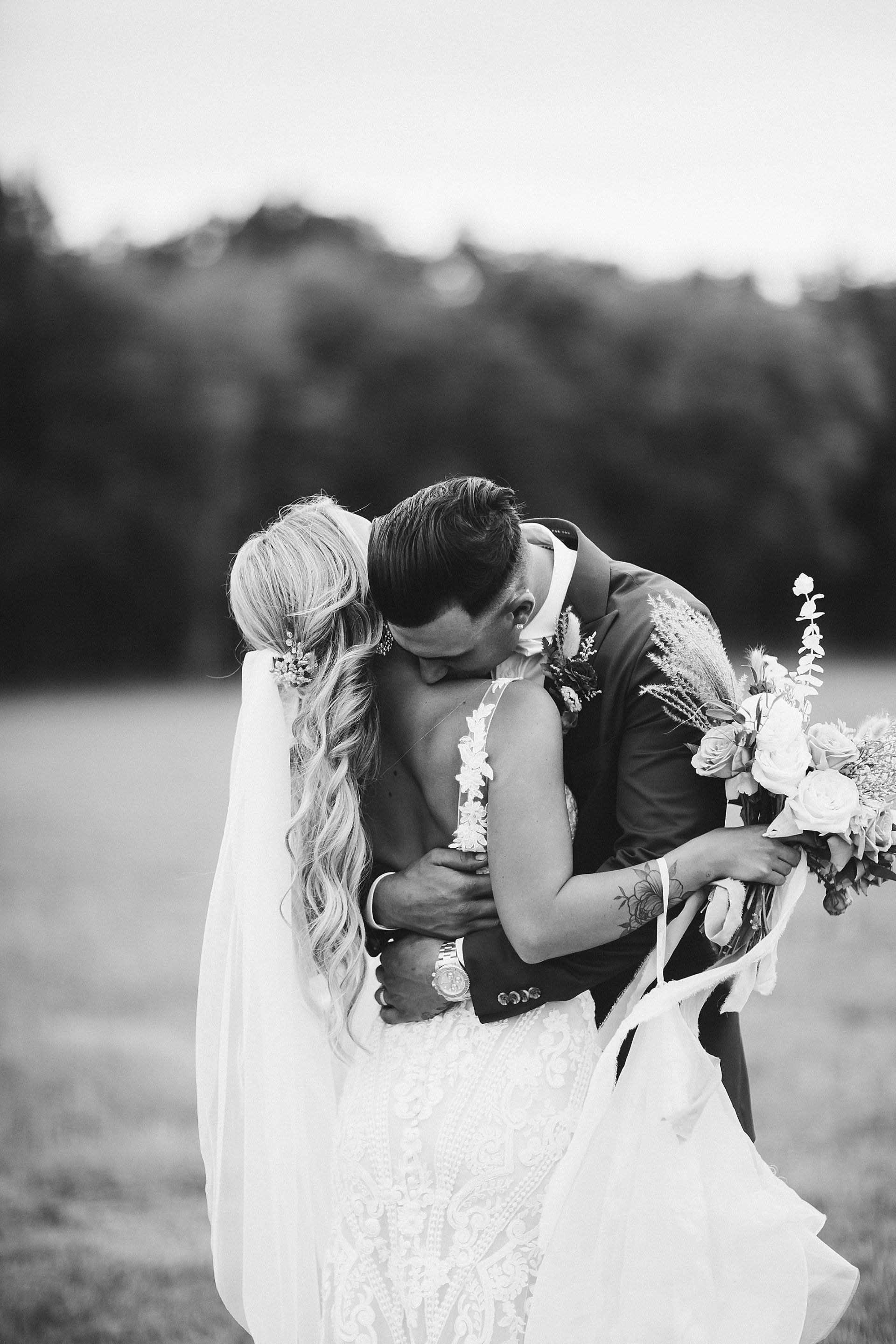 Groom hugs bride, kissing her neck, outdoors. Bride wears a low-back dress and veil, holding flowers.