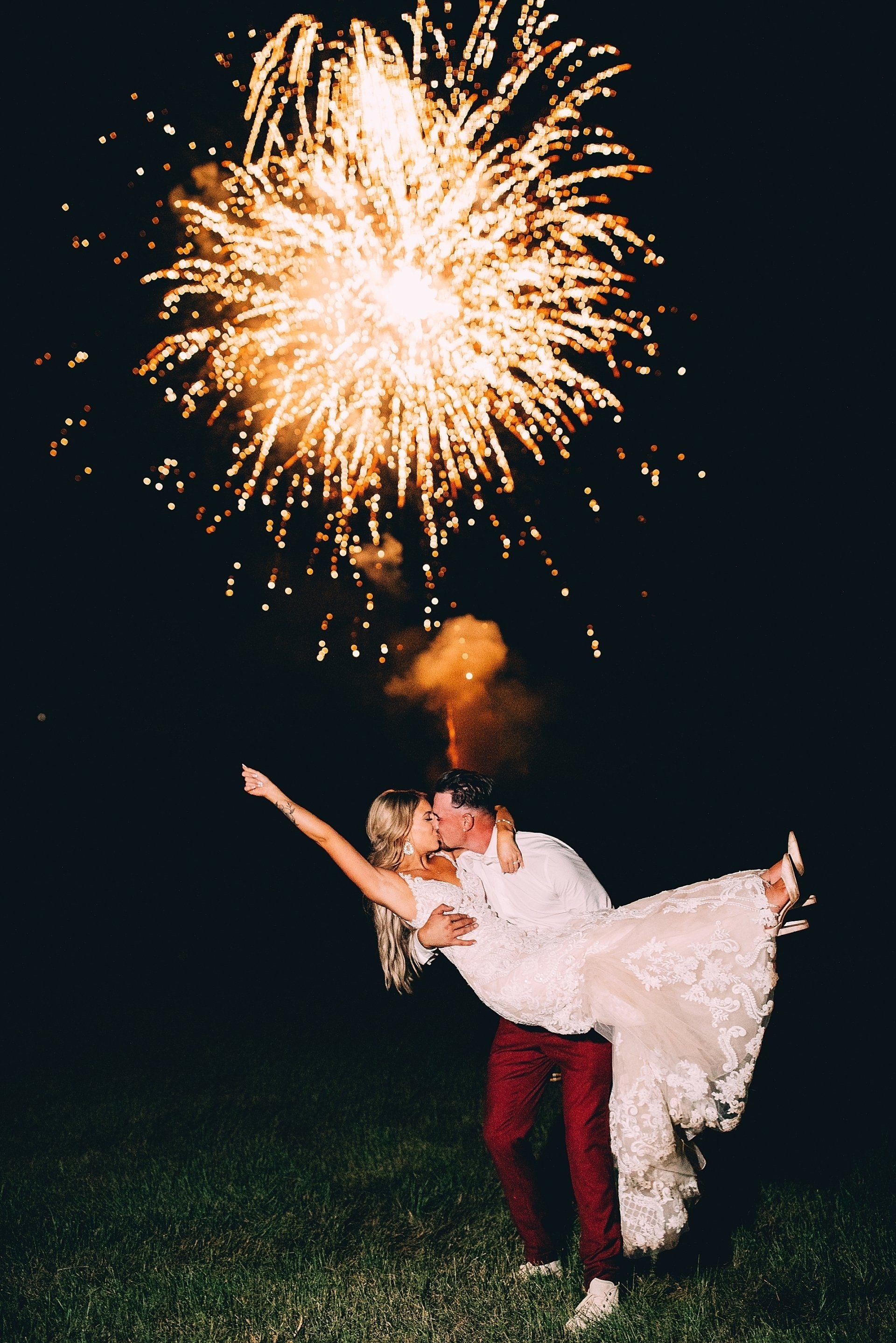 Couple kissing, fireworks exploding overhead. Groom in burgundy pants, bride in white dress, arms outstretched. Night setting.