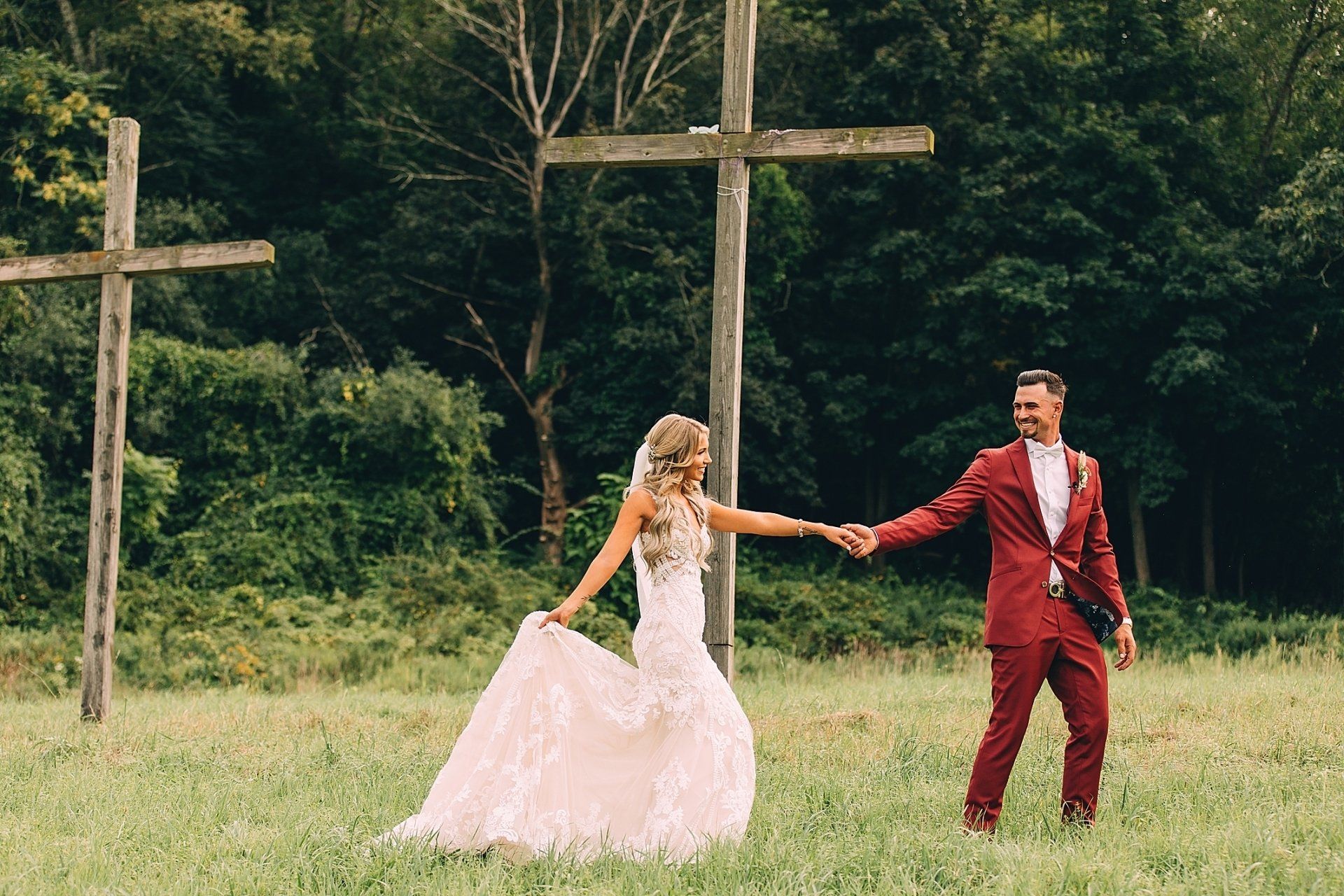 Bride and groom holding hands, walking in a field with wooden crosses in the background.