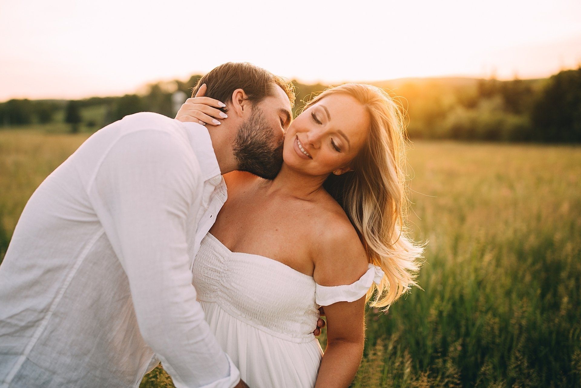 Couple embracing in a field at sunset; man kissing woman's neck, both wearing white. Hudson Valley