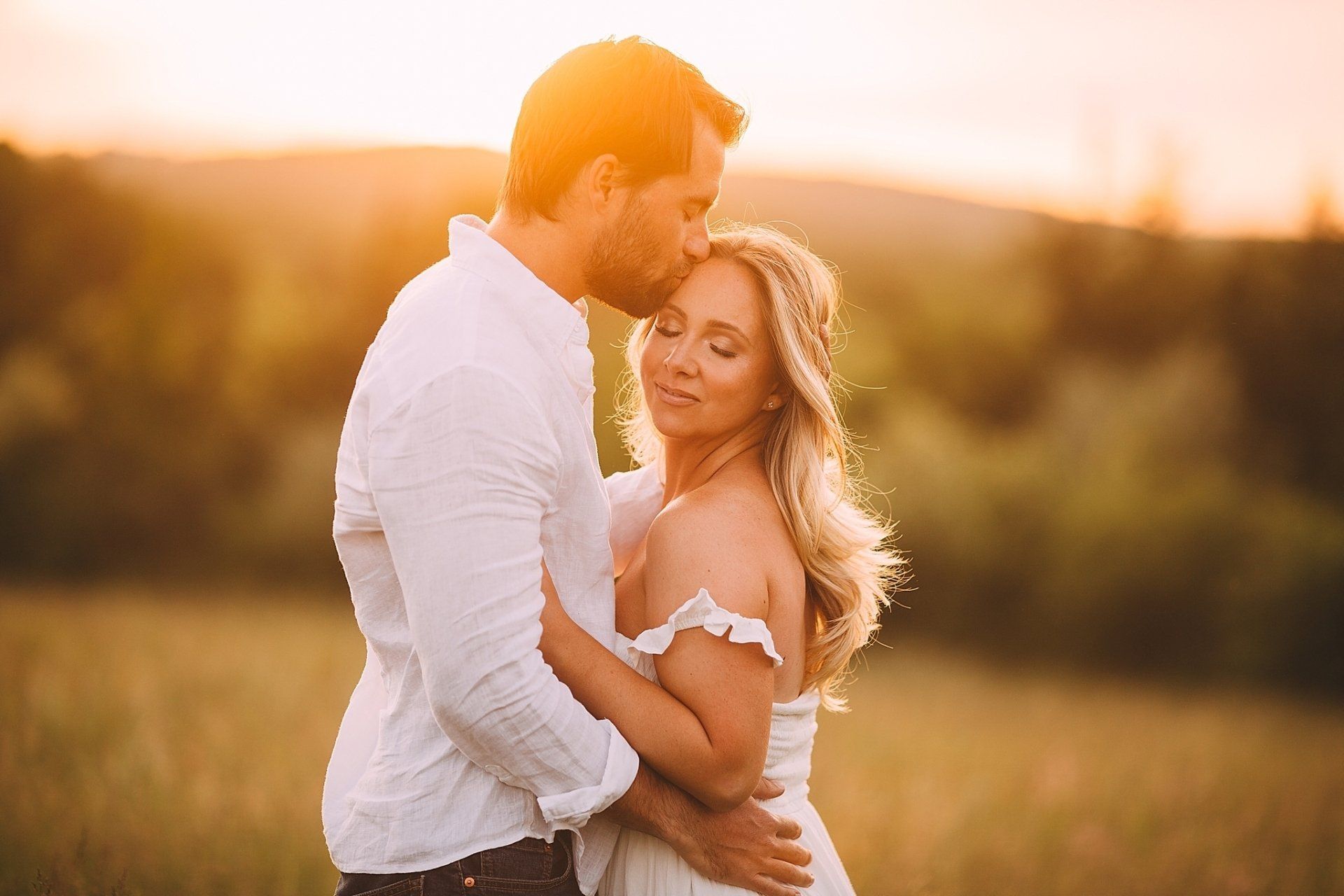 Couple embraces in a field at sunset; man kisses woman's forehead, both with eyes closed. Warm light.
