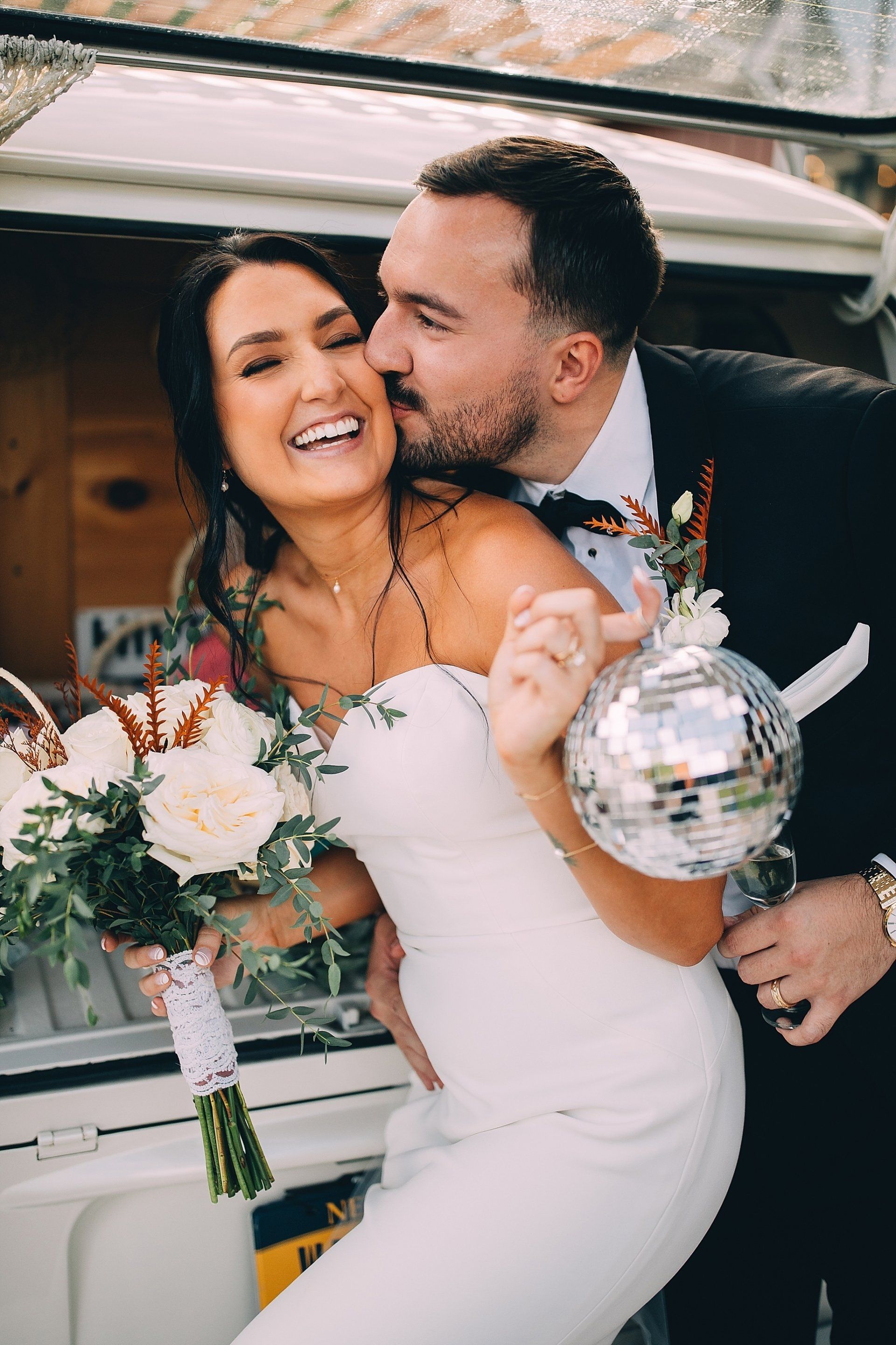 Newlyweds pose; man kisses woman's cheek, holding a disco ball and flowers.