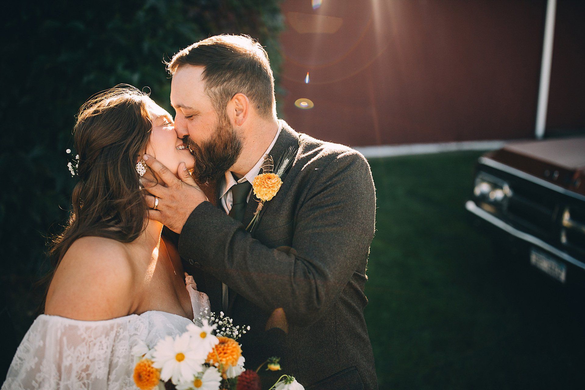 Couple kissing outdoors at their wedding, with a barn and truck in the background.