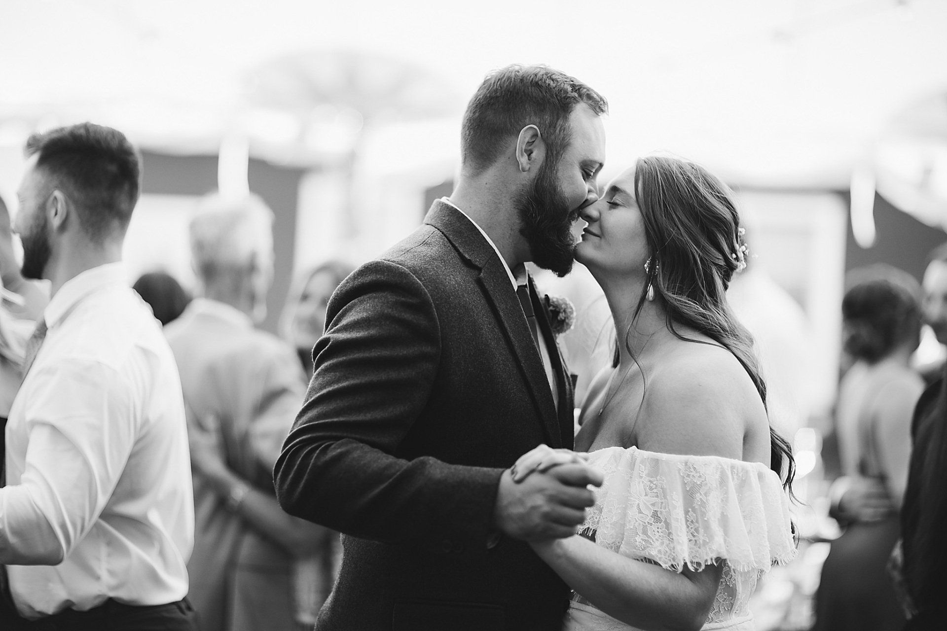 Couple kissing while dancing at a wedding reception; guests in the background. Black and white.